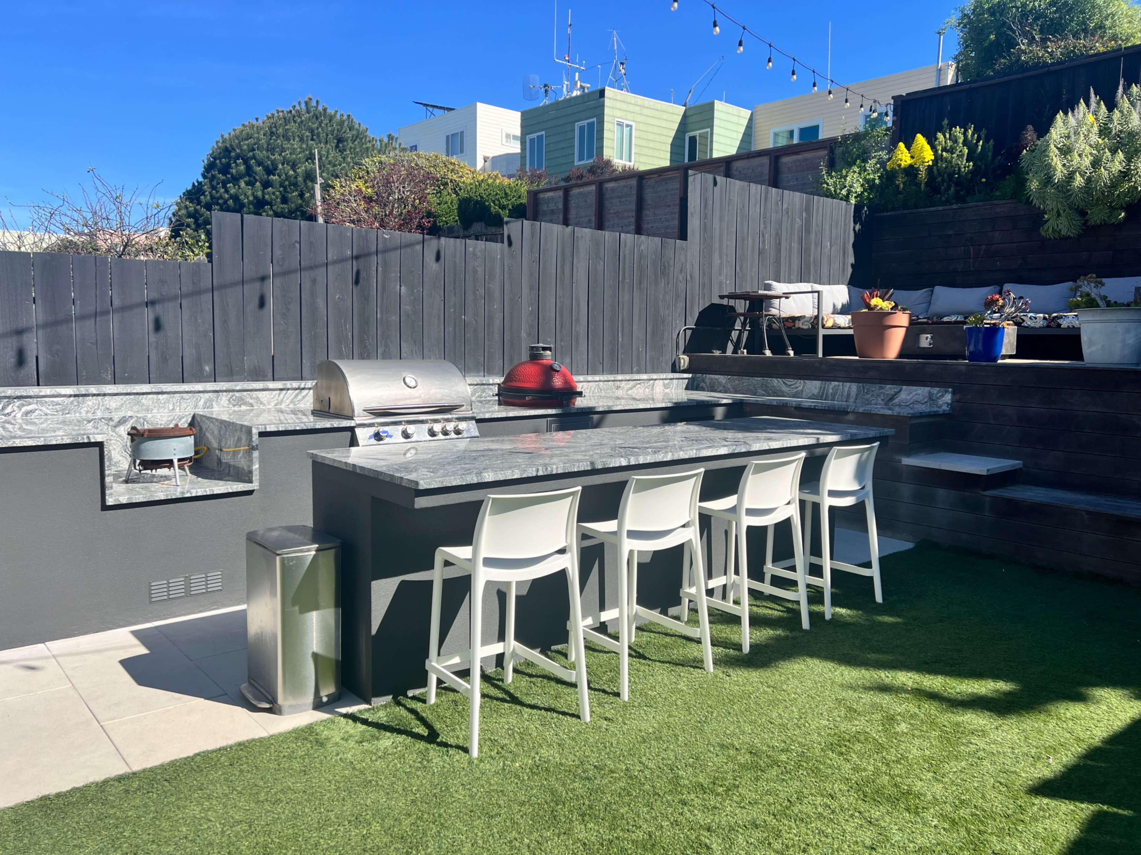 The image shows an outdoor kitchen area featuring a long island with a grill, several bar stools, and a landscaped backdrop with potted plants and a green lawn.