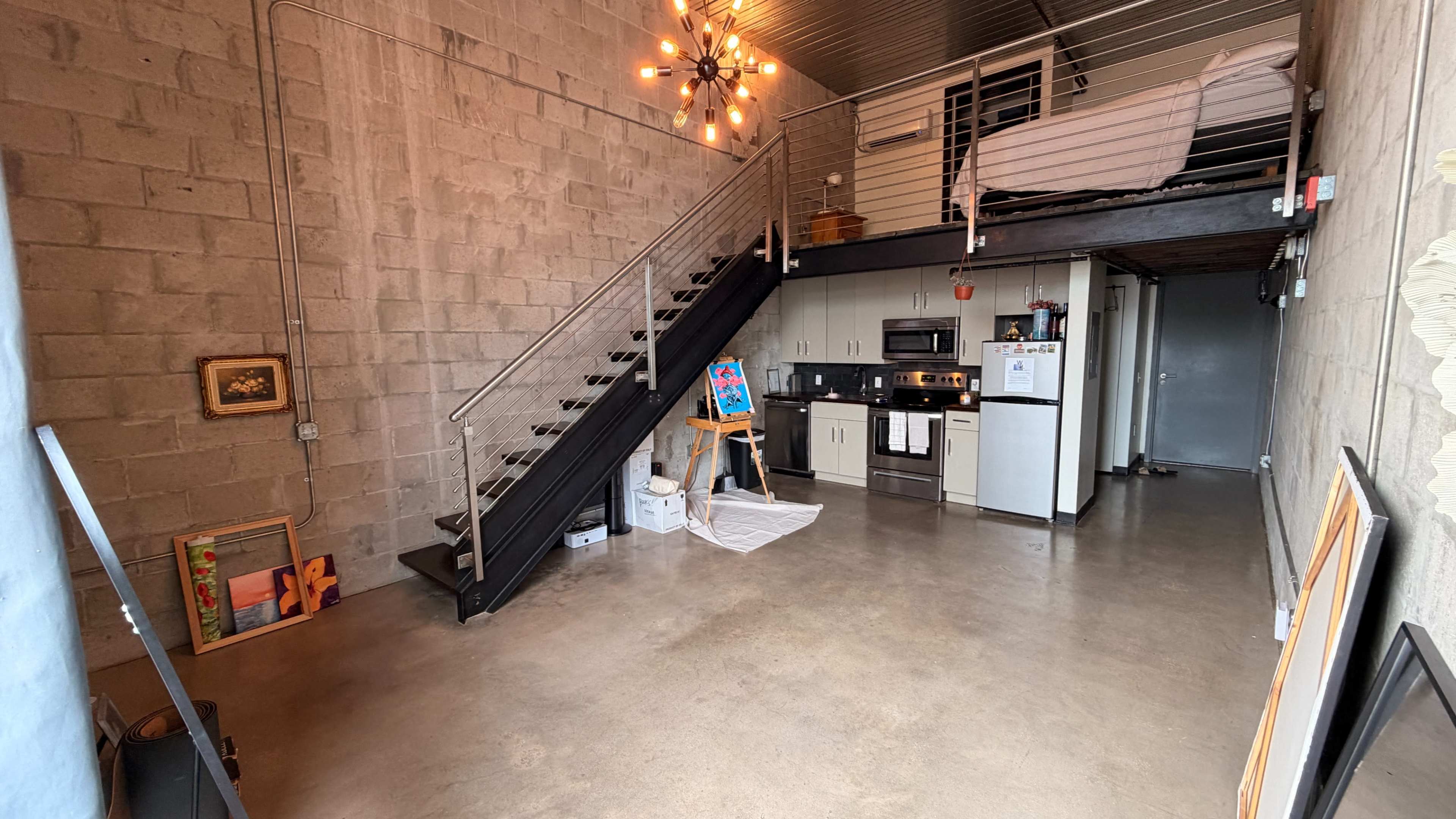 The image shows a modern loft-style interior featuring a kitchen area, a staircase leading to a sleeping loft, and concrete walls.