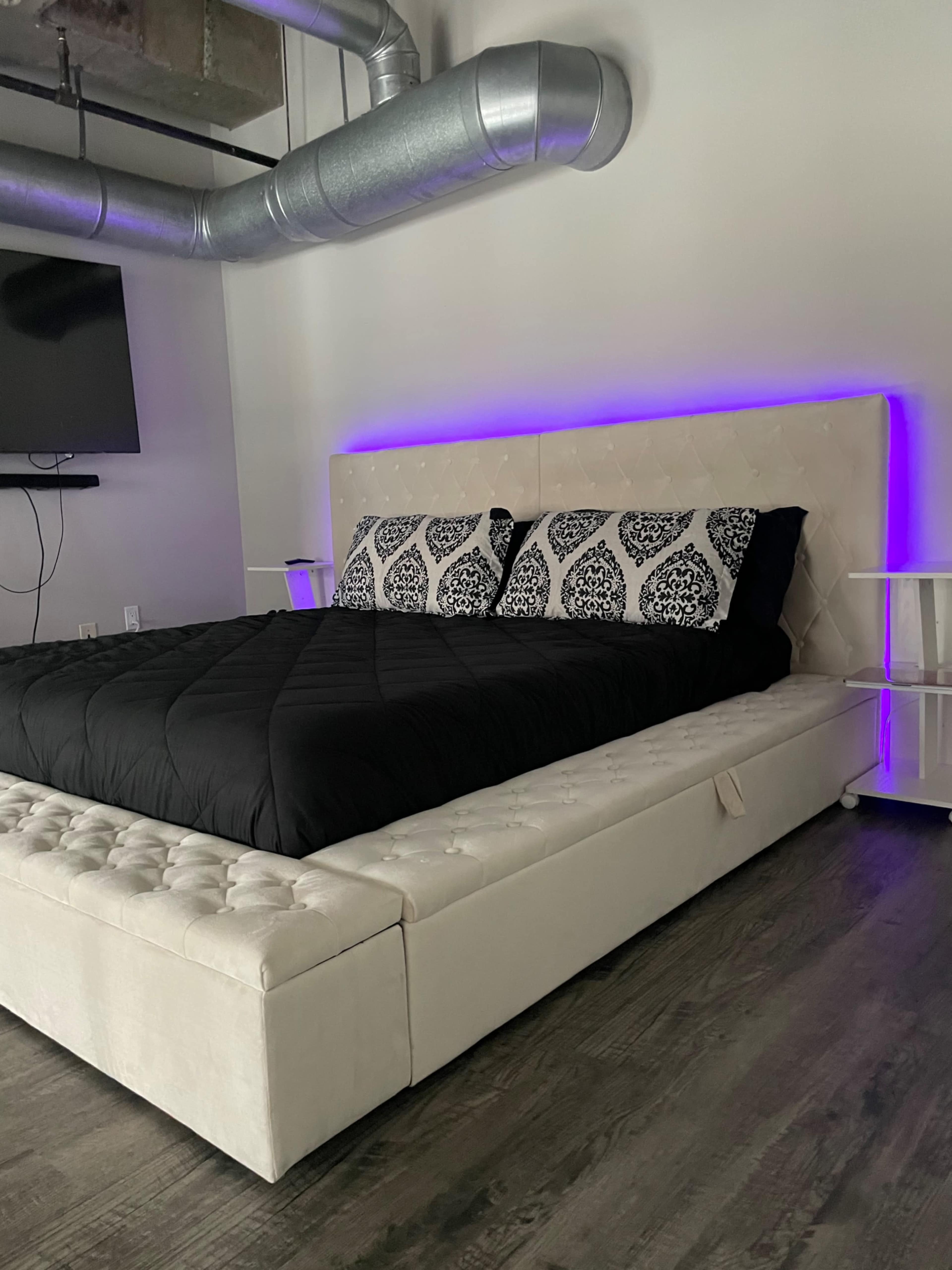A modern bedroom featuring a white tufted bed with a black blanket and decorative pillows, complemented by a purple LED light and a wall-mounted television.