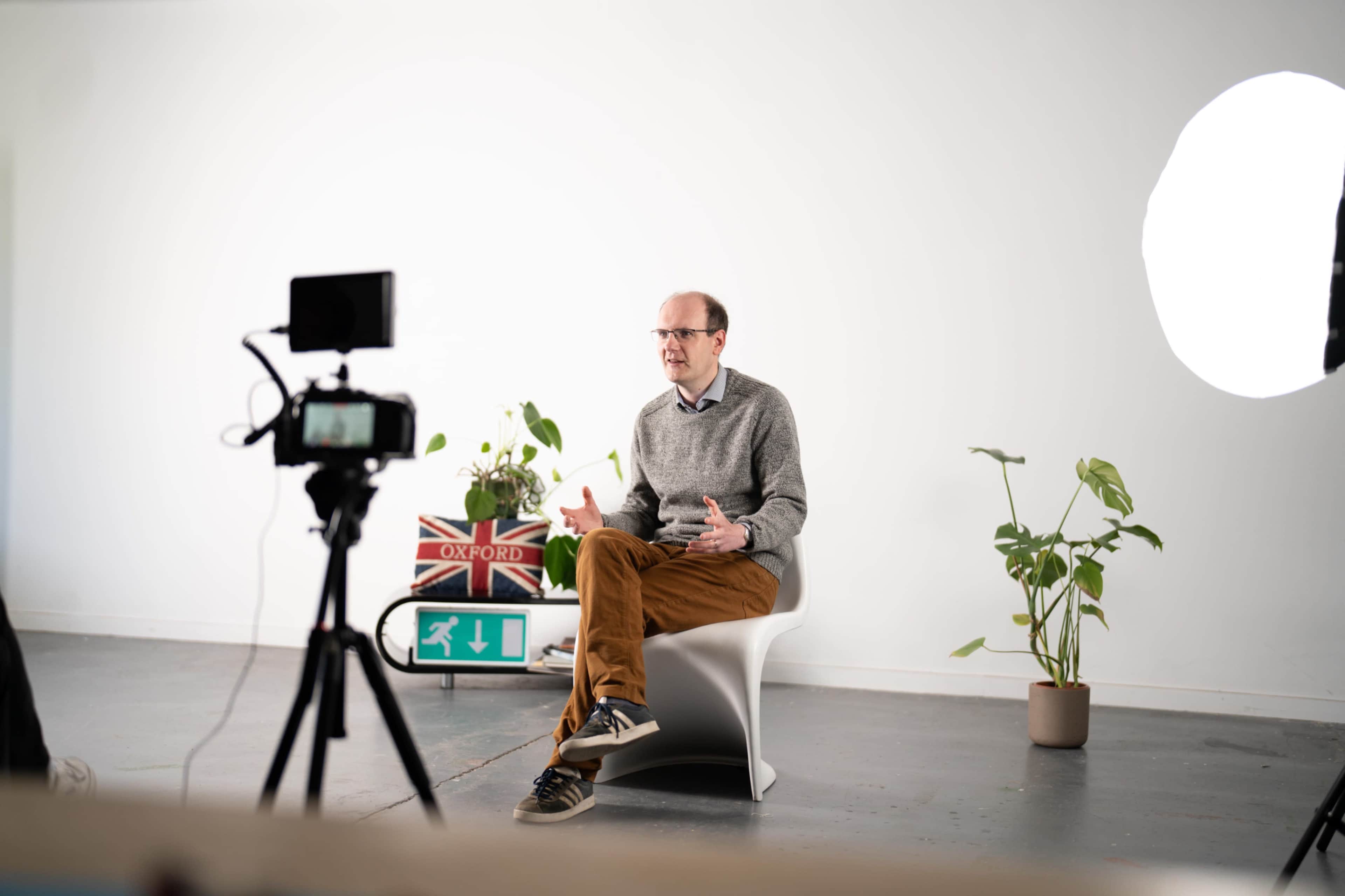 A man in a gray sweater and brown pants sits on a modern chair in front of a camera, speaking in a well-lit indoor setting with plants and a decorative sign in the background.