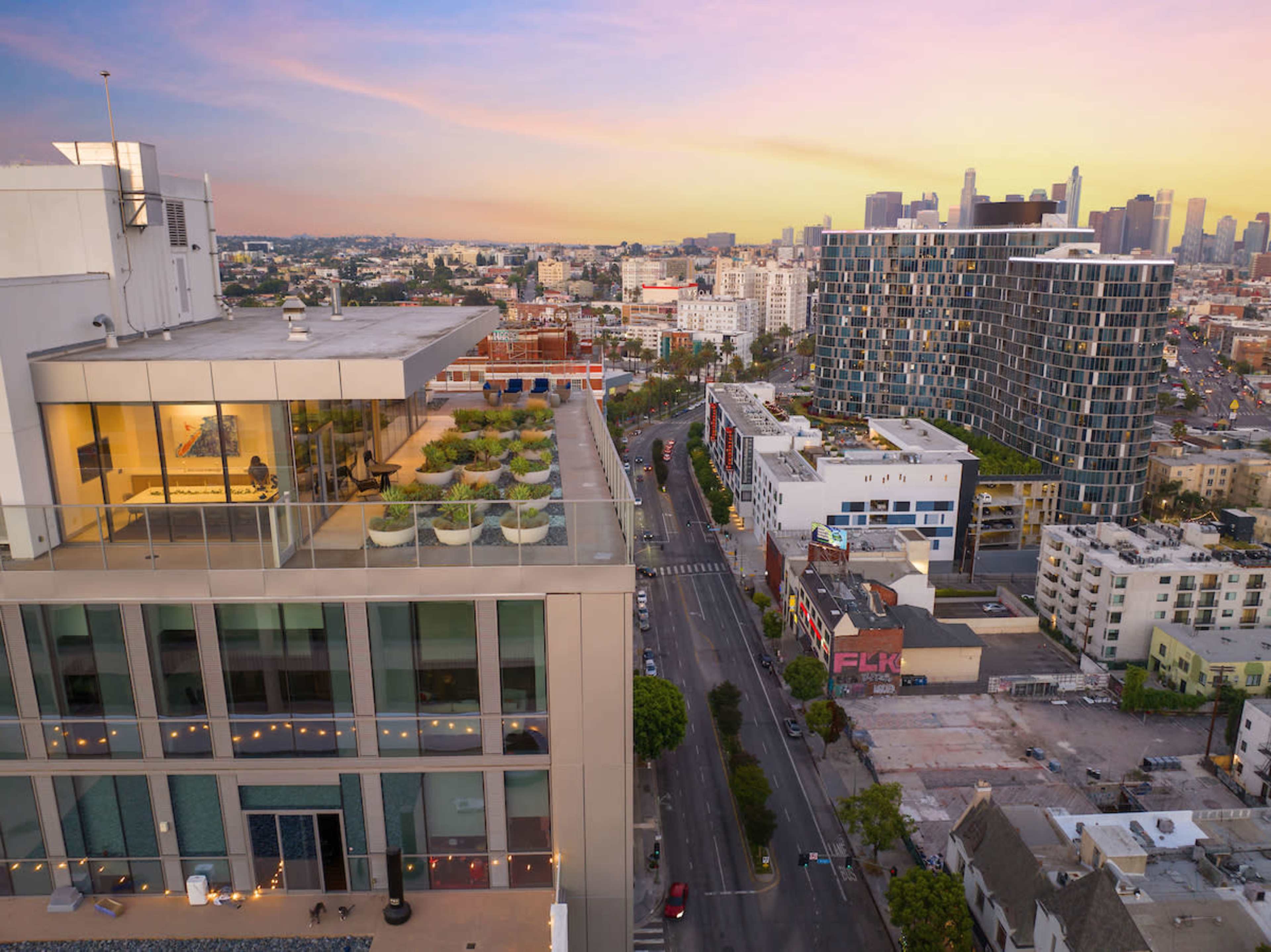 A cityscape at sunset shows a high-rise building with rooftop gardens and an urban street lined with buildings, leading towards a skyline of downtown Los Angeles.