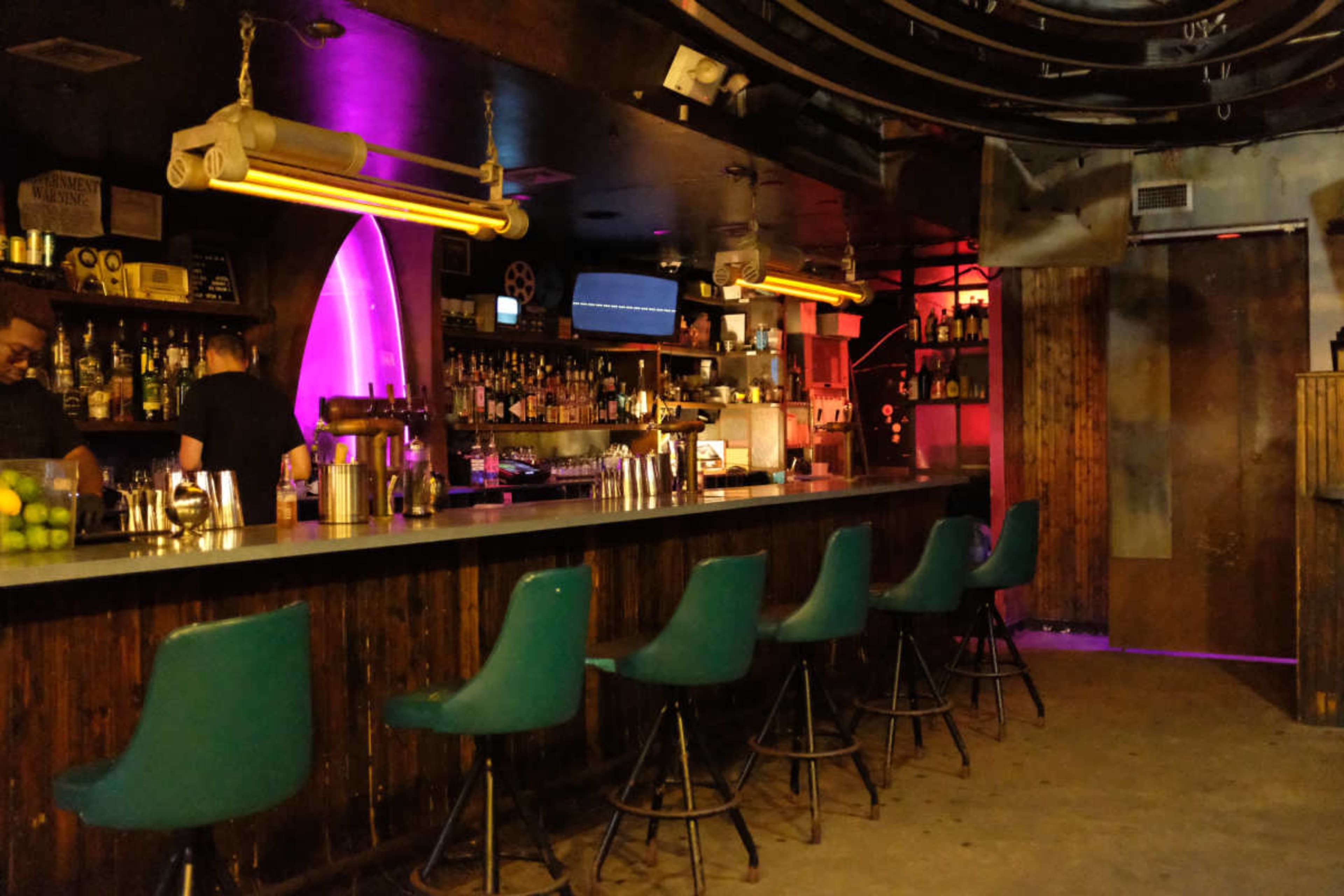 The image shows an empty bar area with green stools lining a wooden counter, illuminated by colorful lights and shelves stocked with bottles.