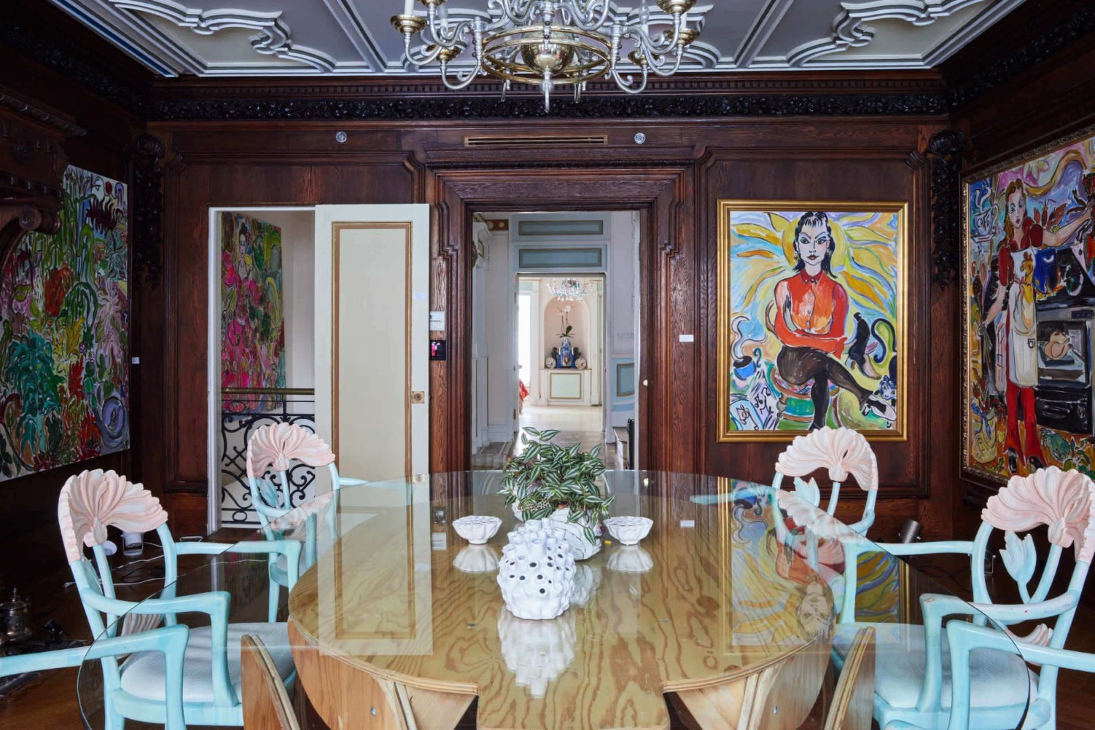 A dining room features a large wooden table surrounded by light blue chairs, with vibrant artworks adorning the dark wooden walls.