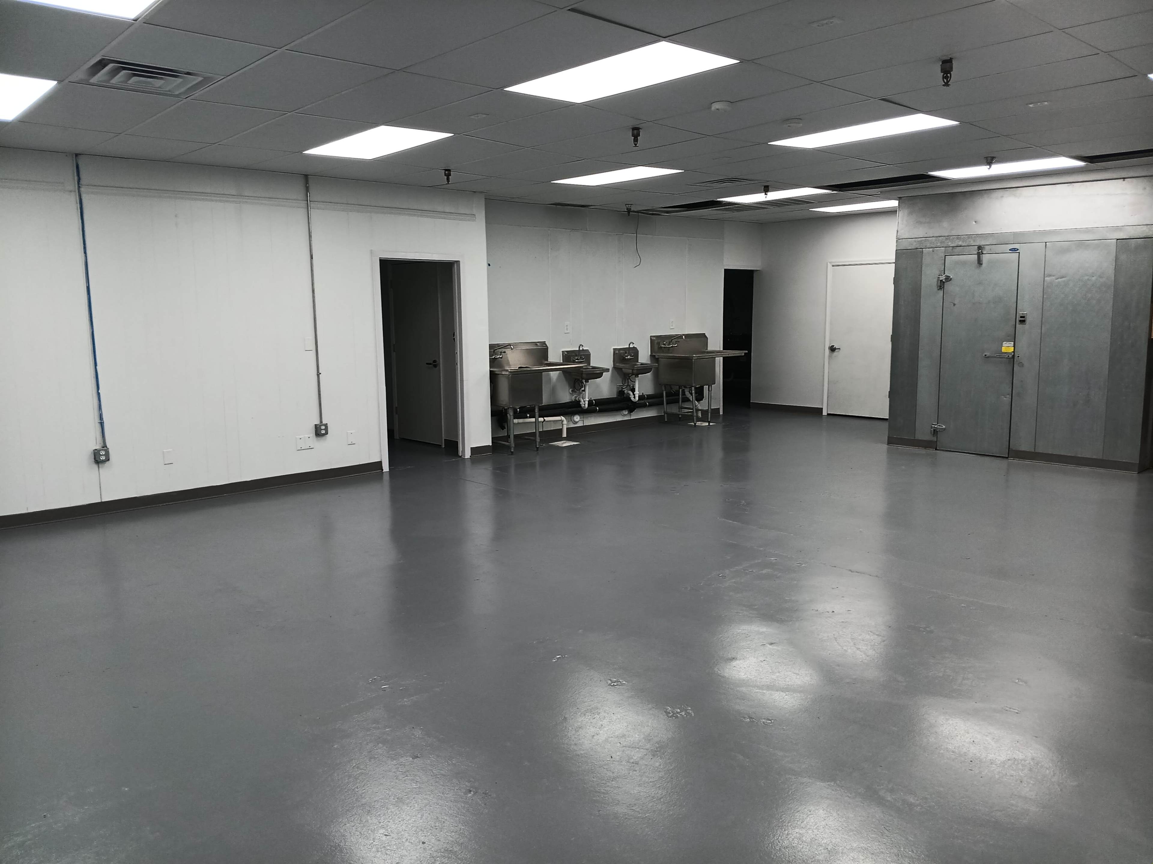 The image shows a large, empty room with gray floors, white walls, and stainless steel sinks against one wall.