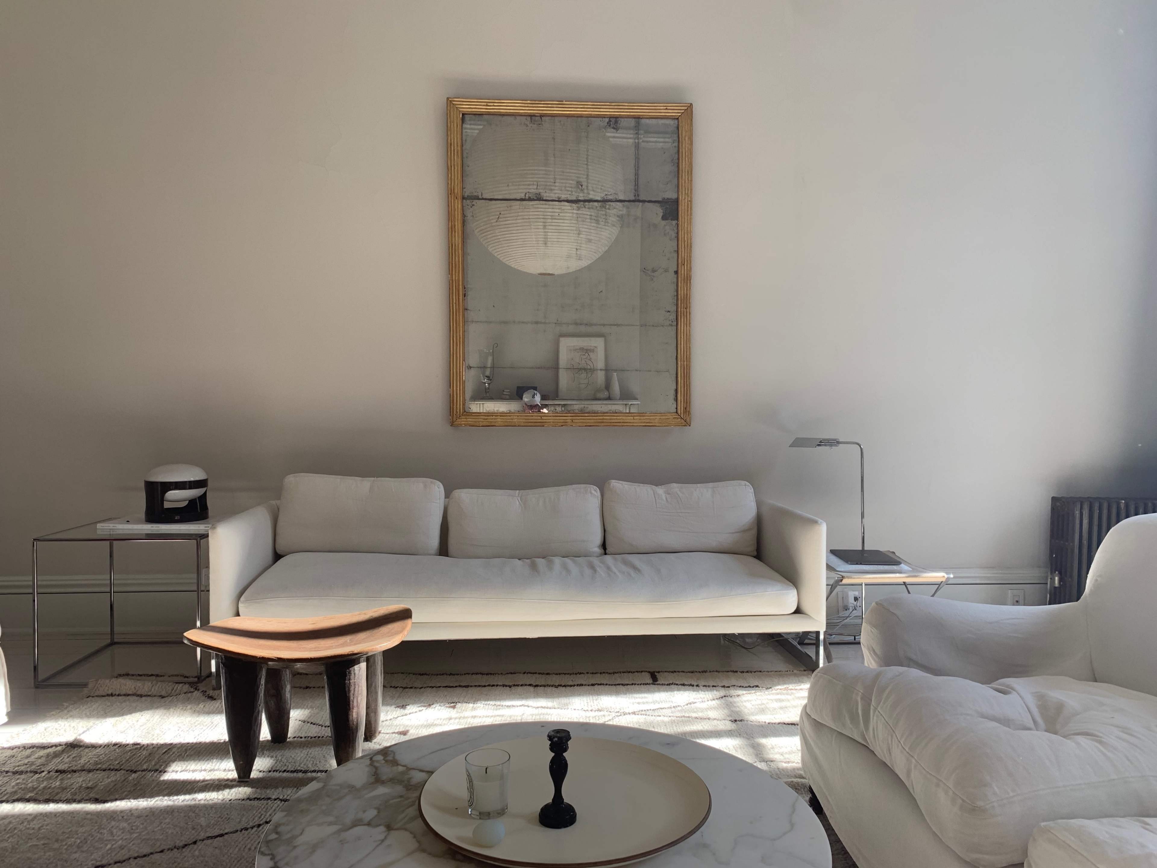 A minimalist living room featuring a white sofa, a round coffee table with a marble top, a small side table, and a large framed mirror on the wall.