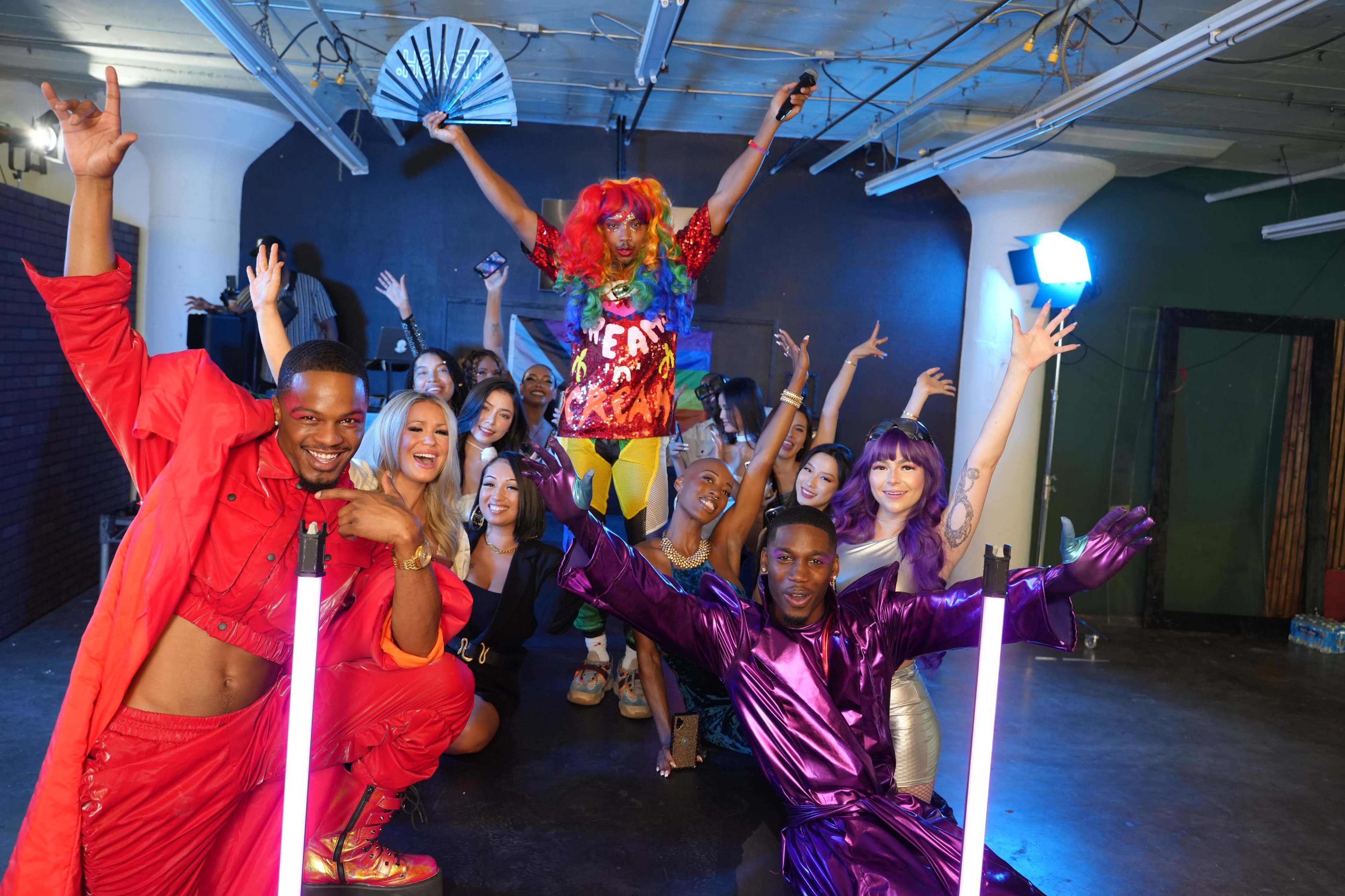 A diverse group of individuals poses energetically in a dimly lit studio, showcasing vibrant outfits and striking poses.