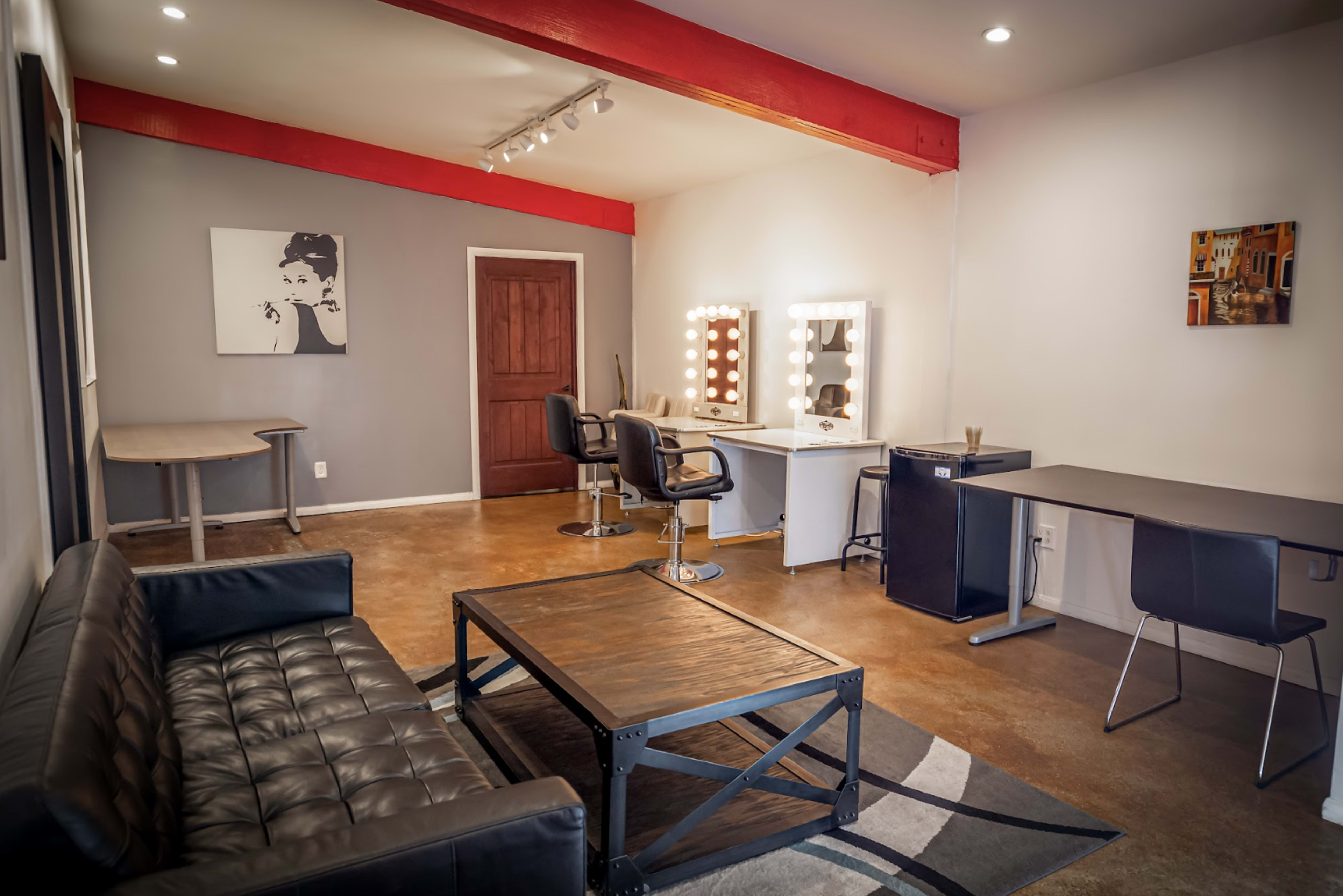 The image shows a well-lit salon or studio space featuring a black sofa, a wooden coffee table, a styling chair, a makeup station with lights, and two work desks.