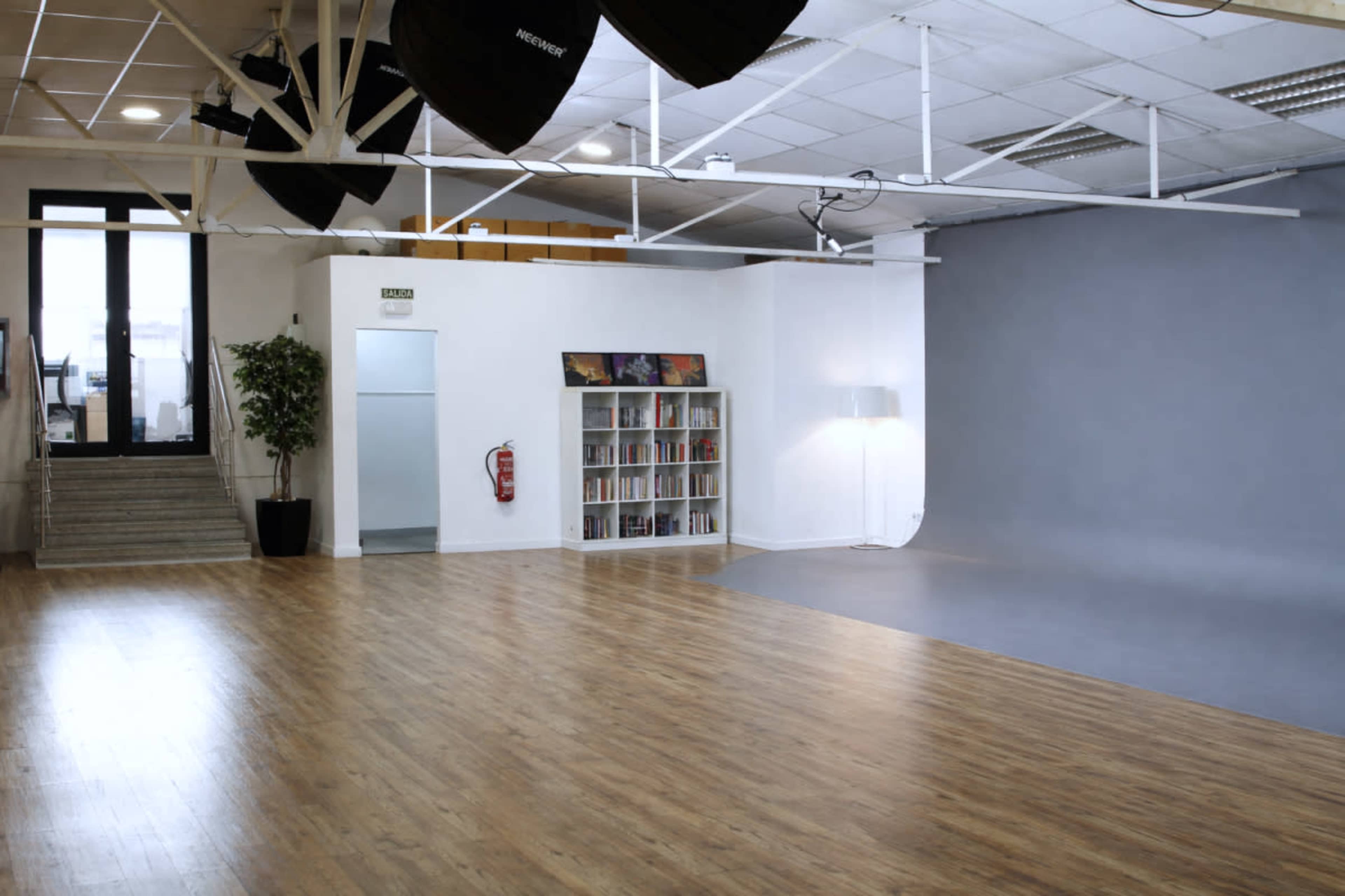 The image shows a spacious photography studio with a wooden floor, white walls, a gray backdrop, and a bookshelf filled with books.