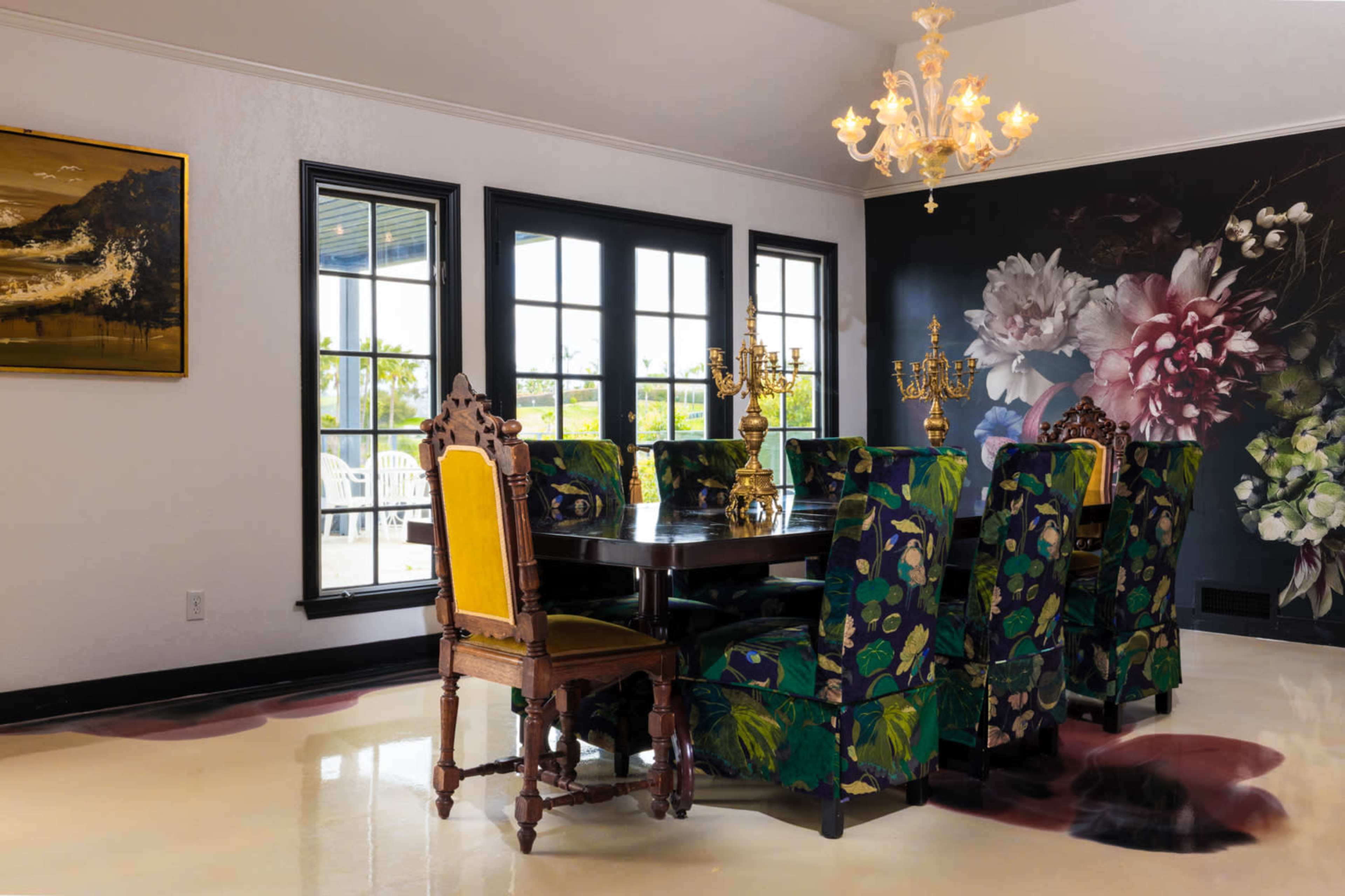 The image shows a dining room featuring a large table surrounded by ornate chairs with floral upholstery, a chandelier overhead, and large windows providing natural light.