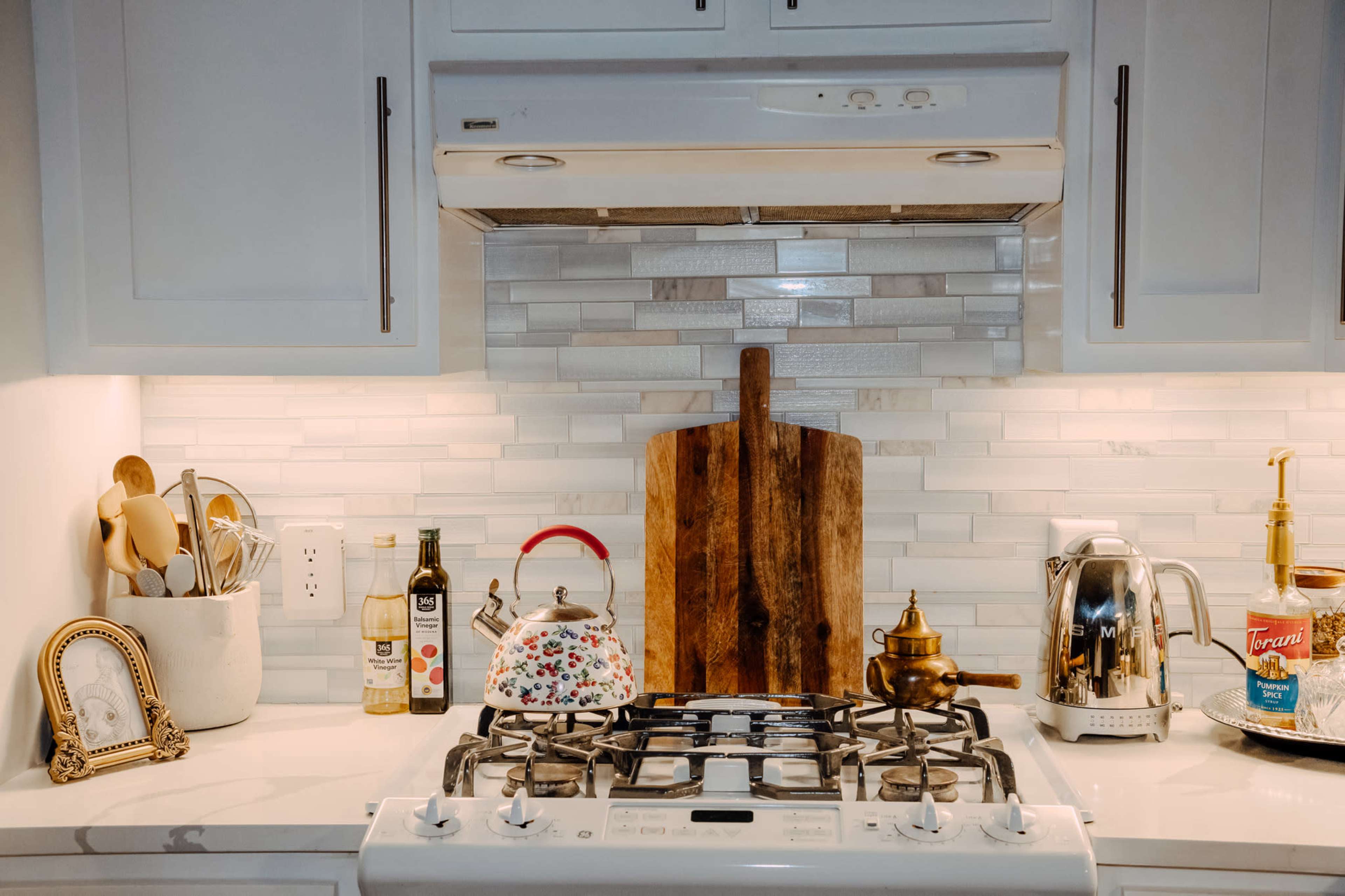 A kitchen countertop features a gas stove, a wooden cutting board, various utensils, and a few decorative items, all against a white tiled backsplash.