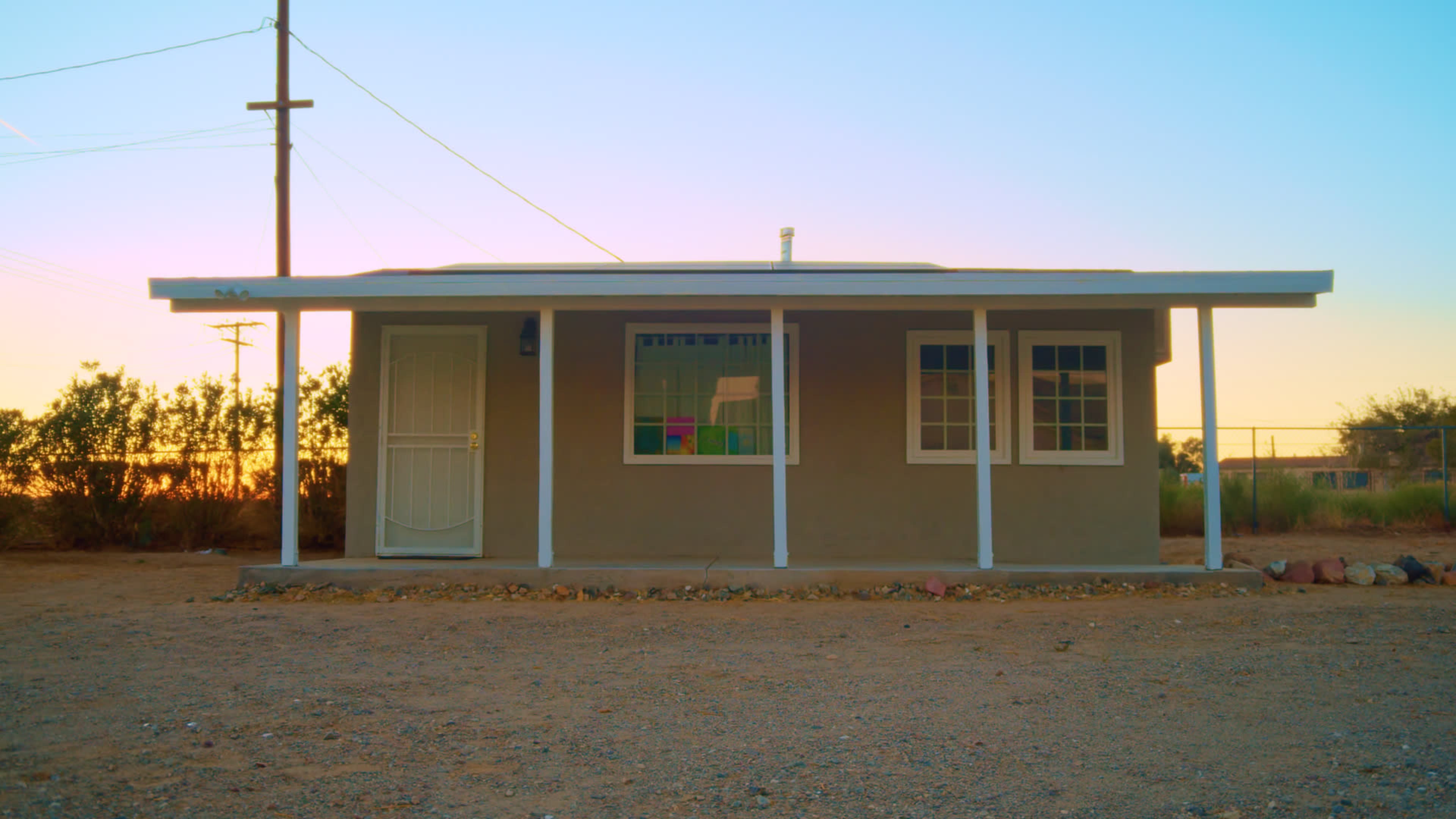 A small, single-story house with a porch is situated in a sparse, sandy landscape under a colorful sunset.