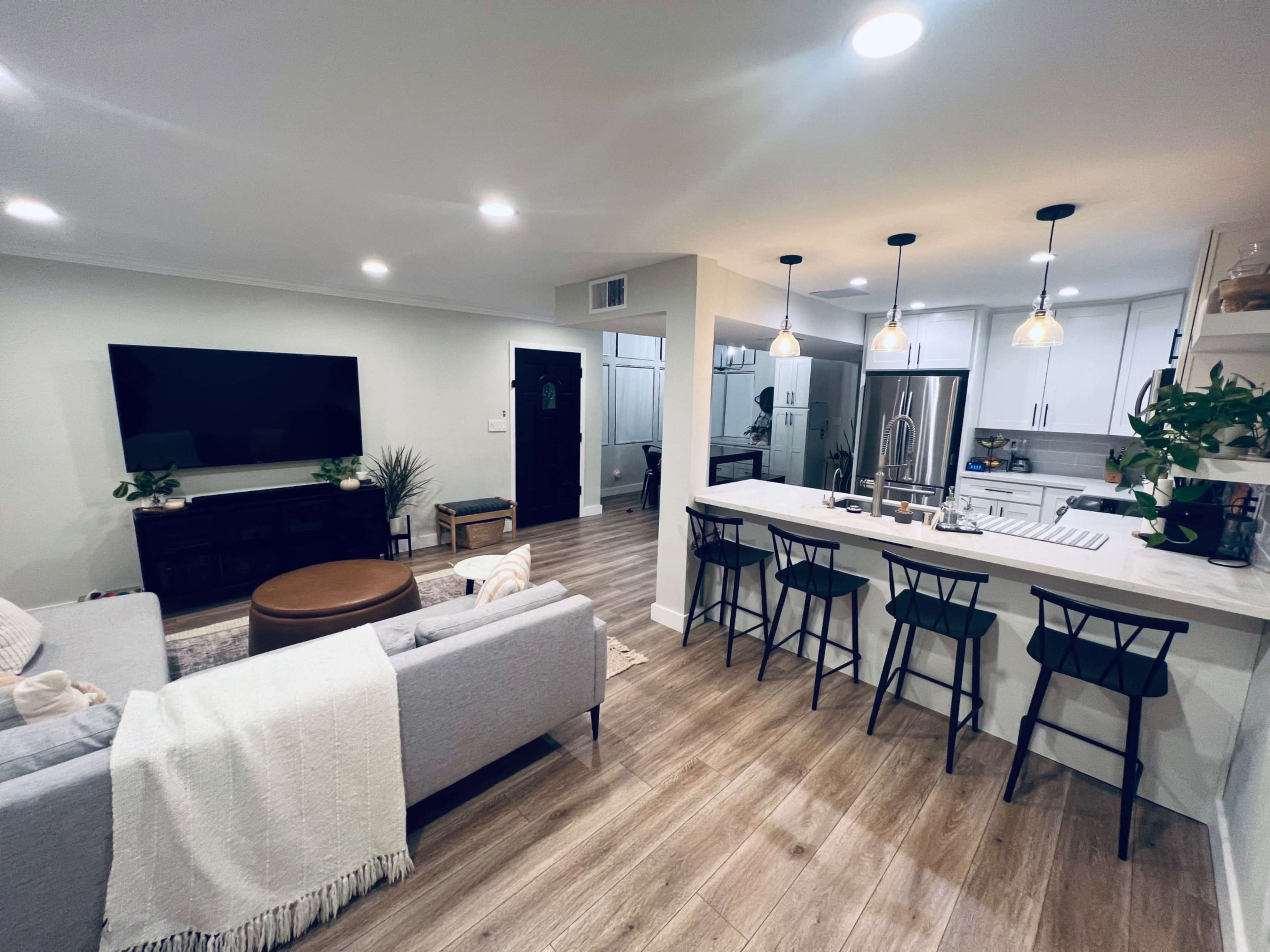 The image shows a modern living room and kitchen area with a gray sofa, a wooden coffee table, a large TV, and a kitchen counter with bar stools.