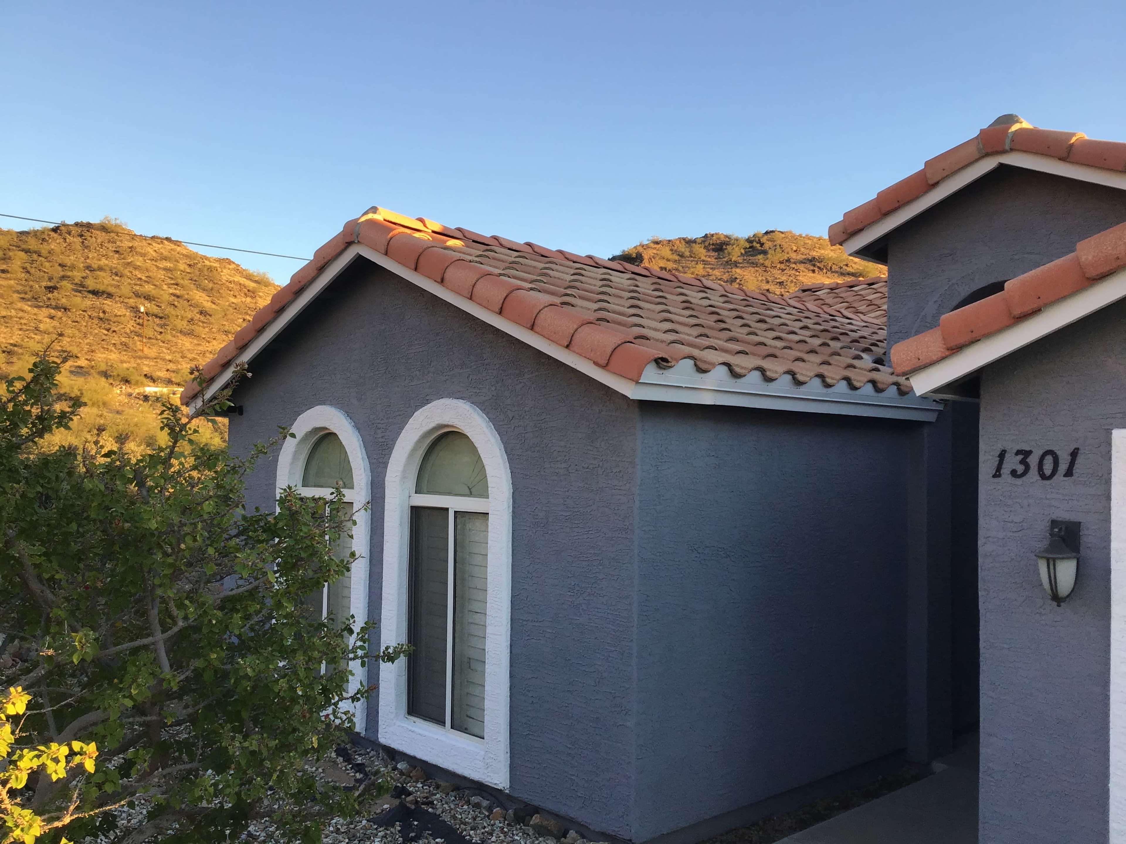 A single-story house with a gray exterior, arched windows, and a tiled roof, situated near a hillside.