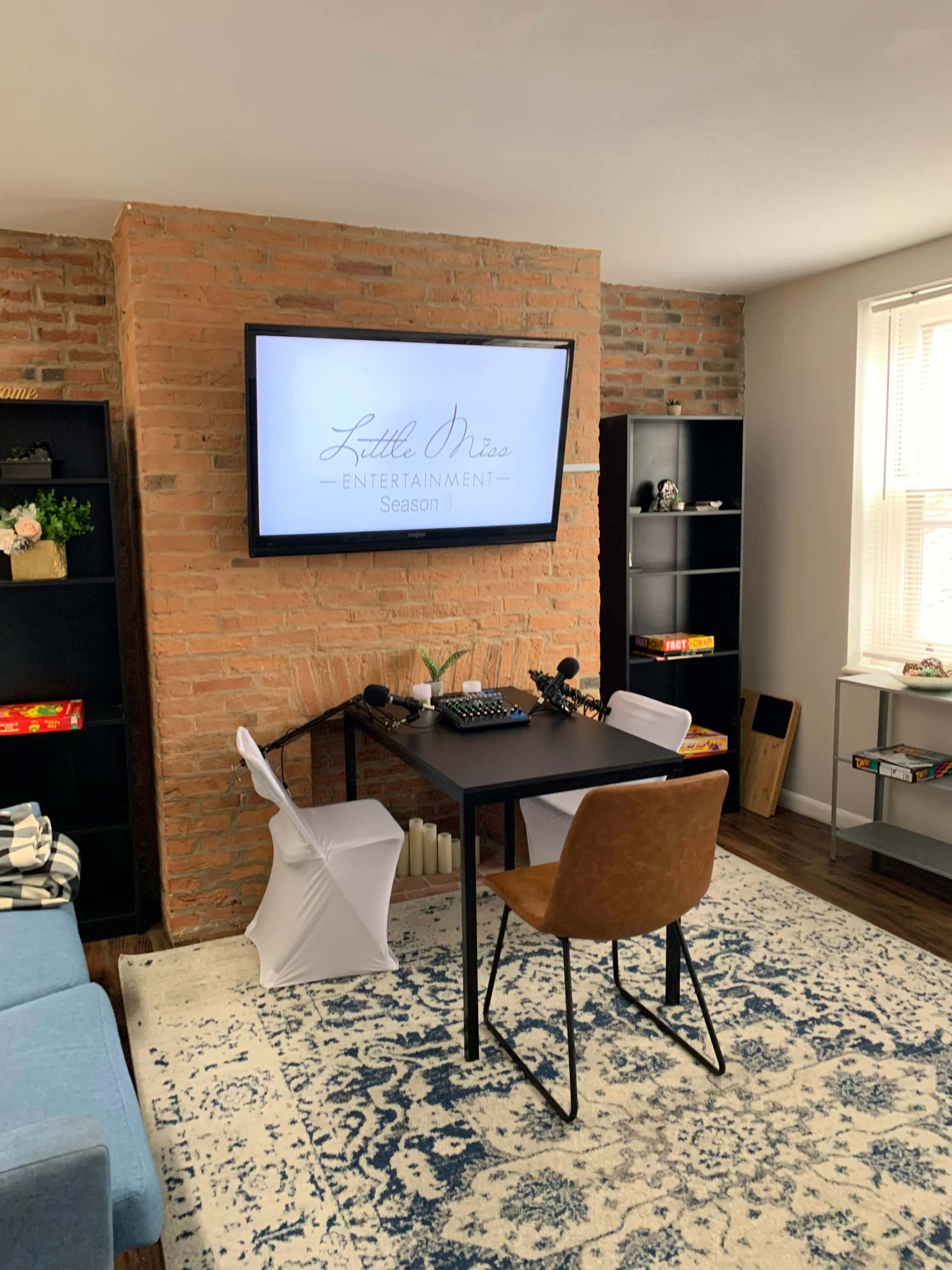 A home office features a black desk with a keyboard and monitor against a brick wall, surrounded by shelves and a light-colored rug.