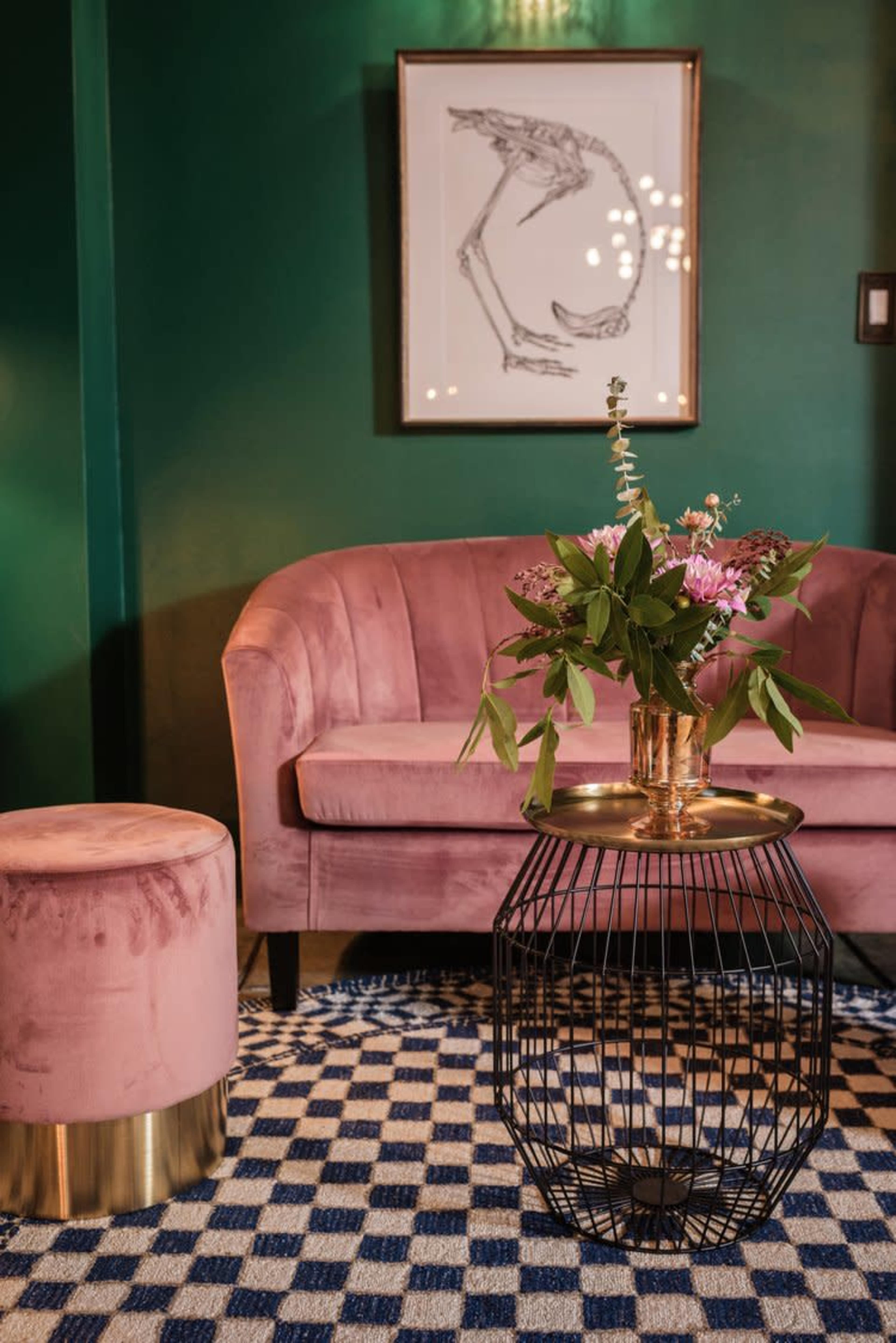A pink upholstered sofa and a round stool are positioned next to a gold-accented wire table, all set against a dark green wall with a decorative floral arrangement on the table.