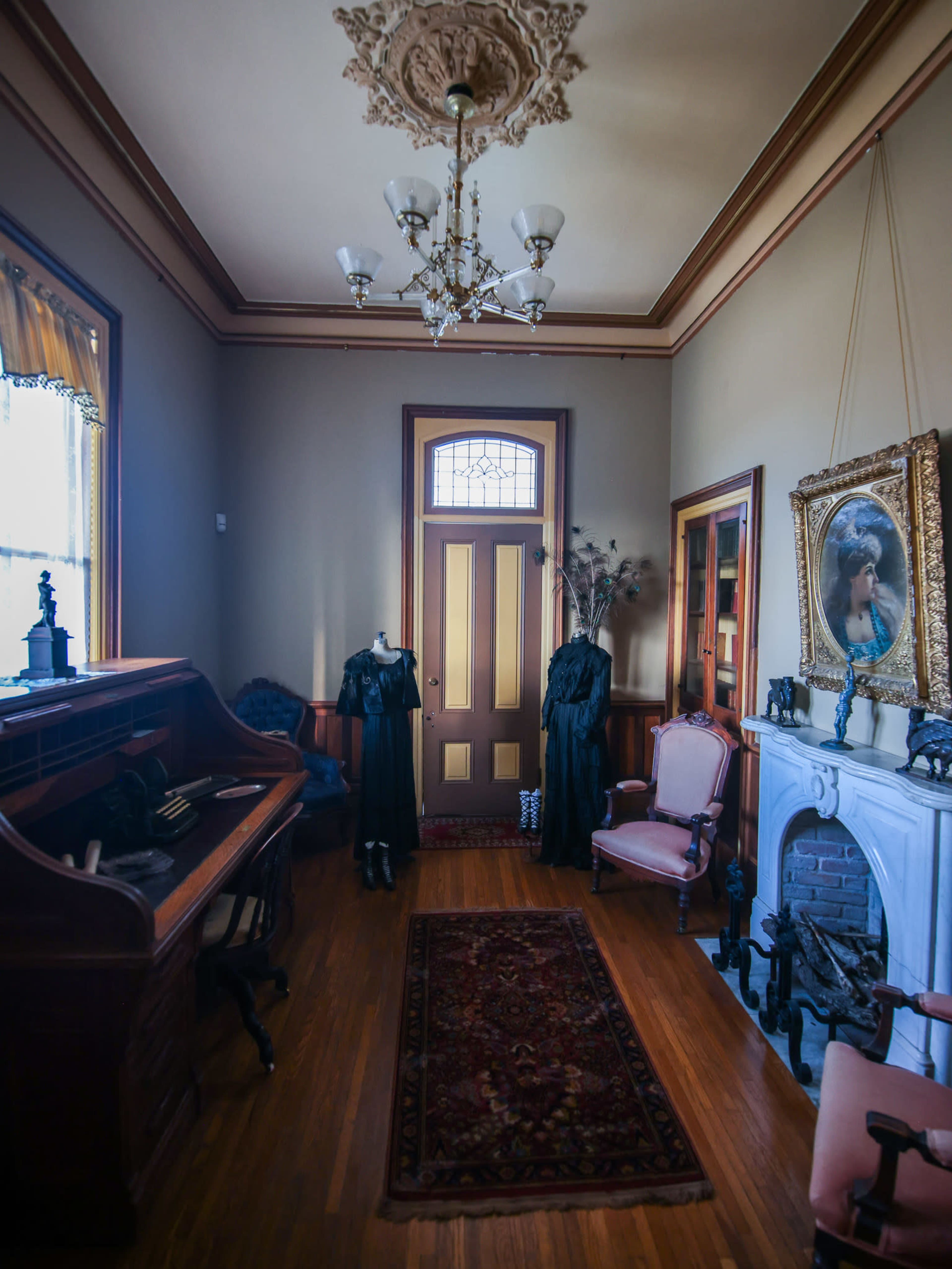A vintage room with a wooden writing desk, two dark dresses on mannequins, pink upholstered chairs, and a large decorative mirror above a fireplace.