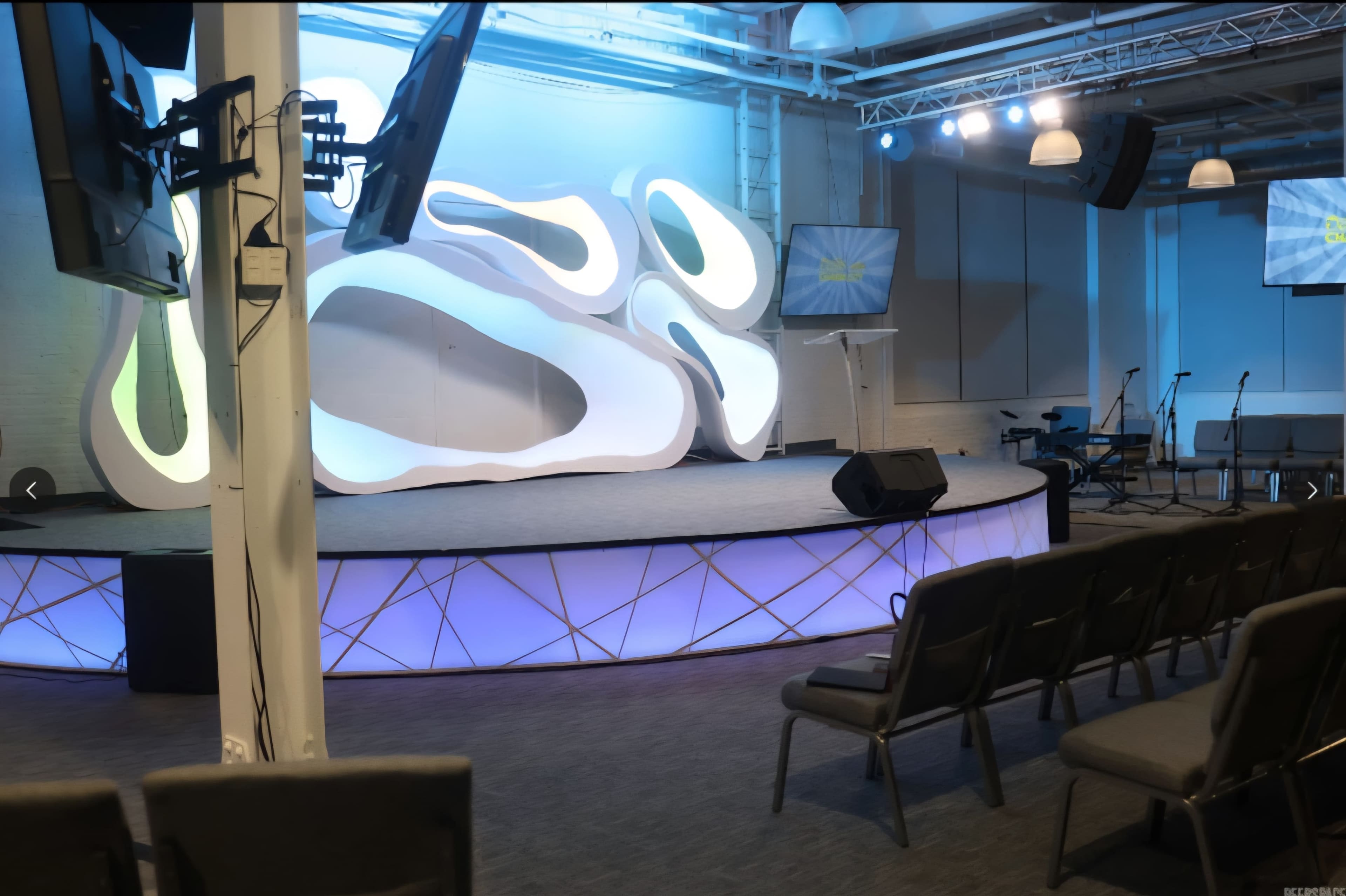 The image shows a modern stage setup in a venue, featuring a curved backdrop and several monitors, with rows of empty chairs facing the stage.