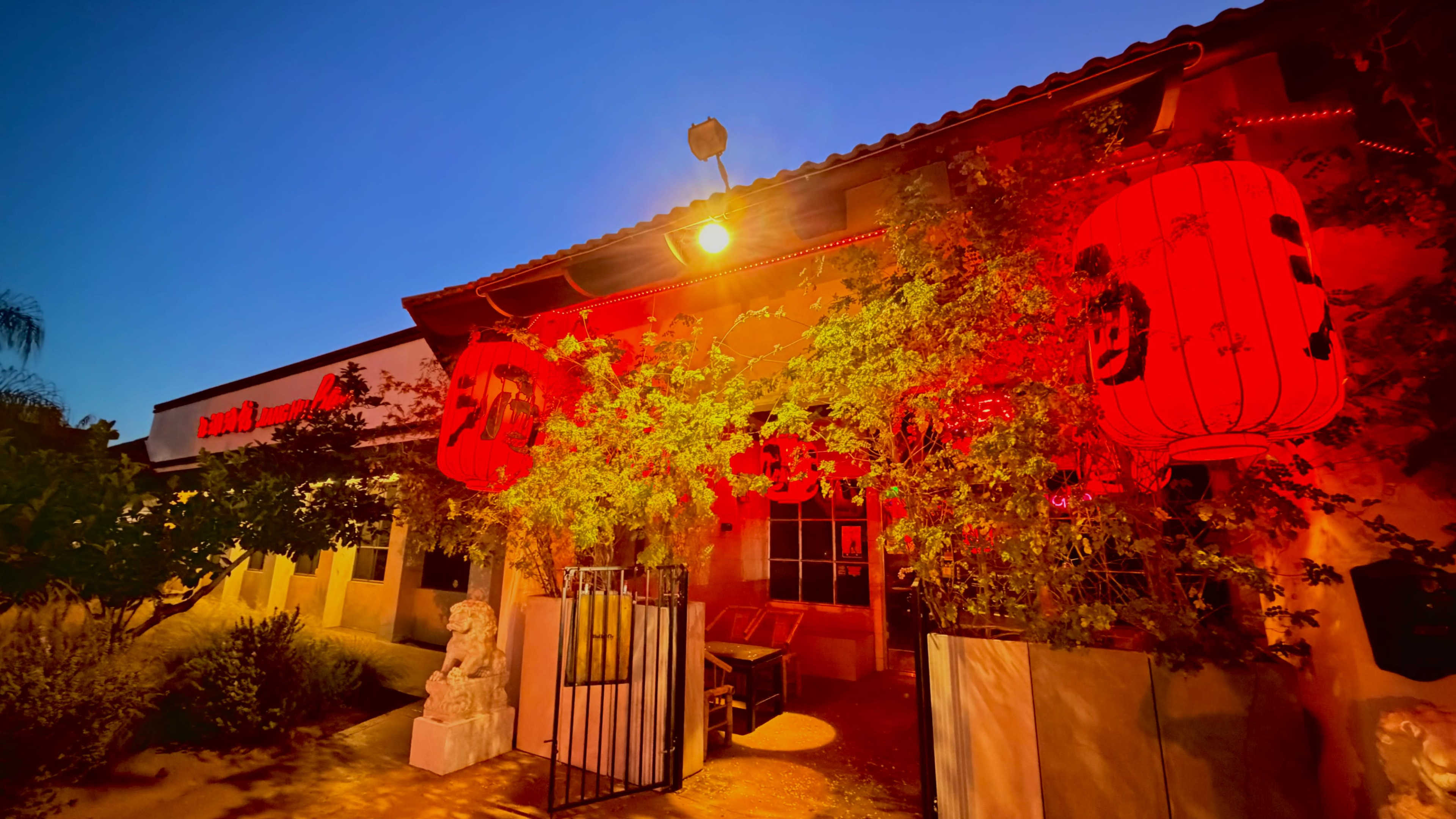 The exterior of a restaurant features red lanterns and vibrant greenery against a twilight sky.