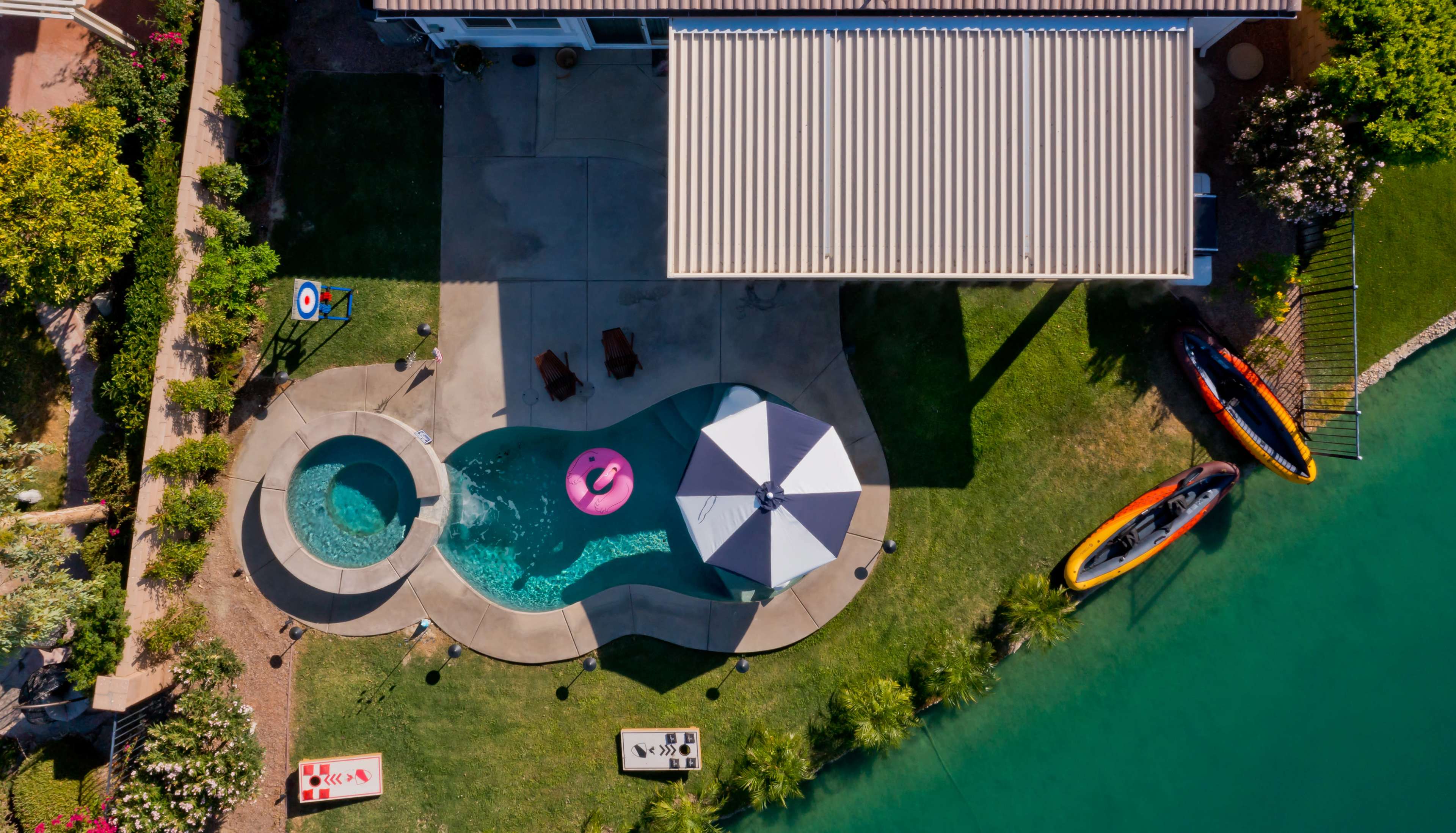 An aerial view of a backyard with a swimming pool, hot tub, patio umbrella, two lounge chairs, and a kayaks by a water body.