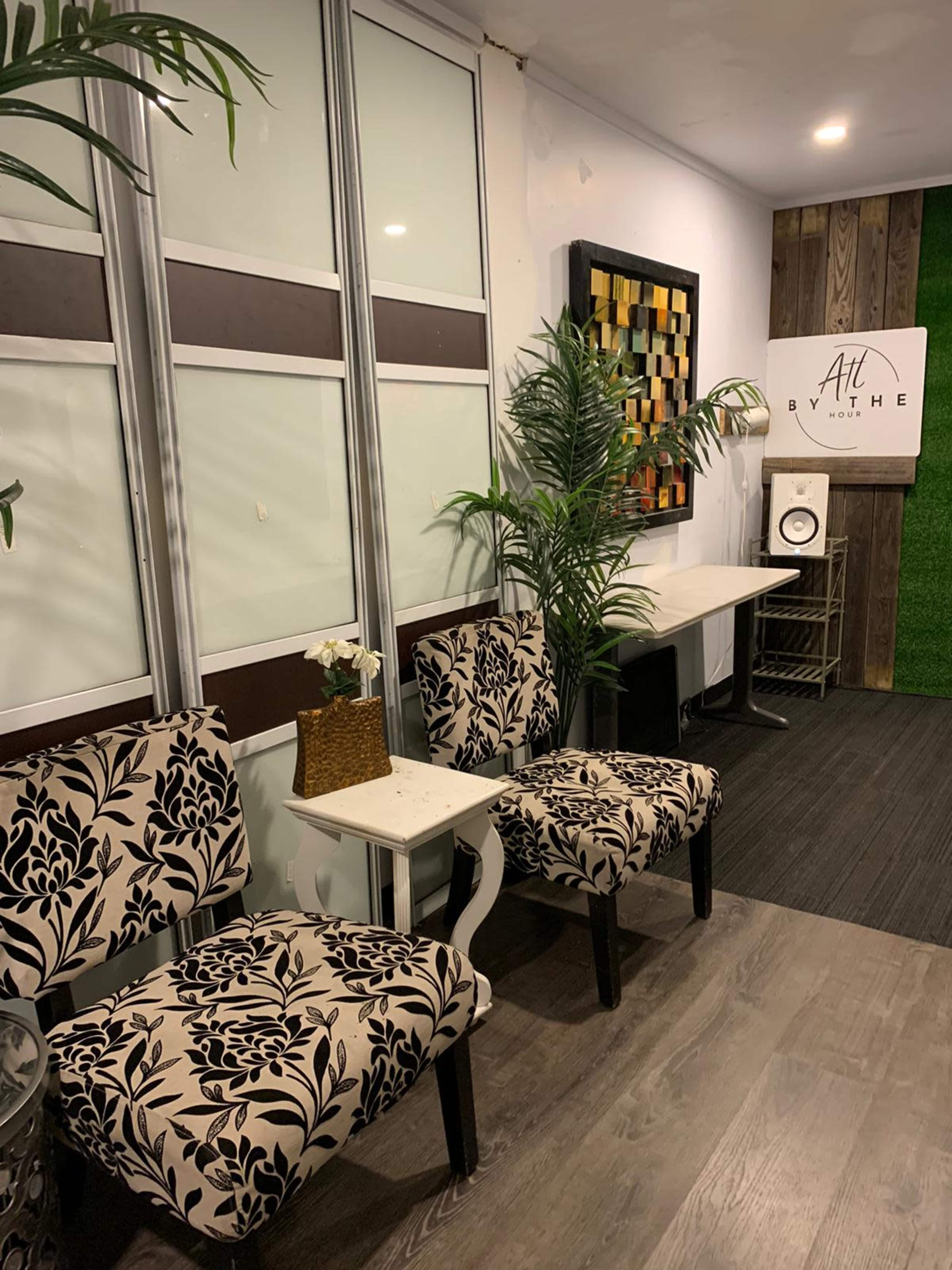 A small waiting area with two patterned chairs, a side table, plants, and a wall featuring art and a sign.
