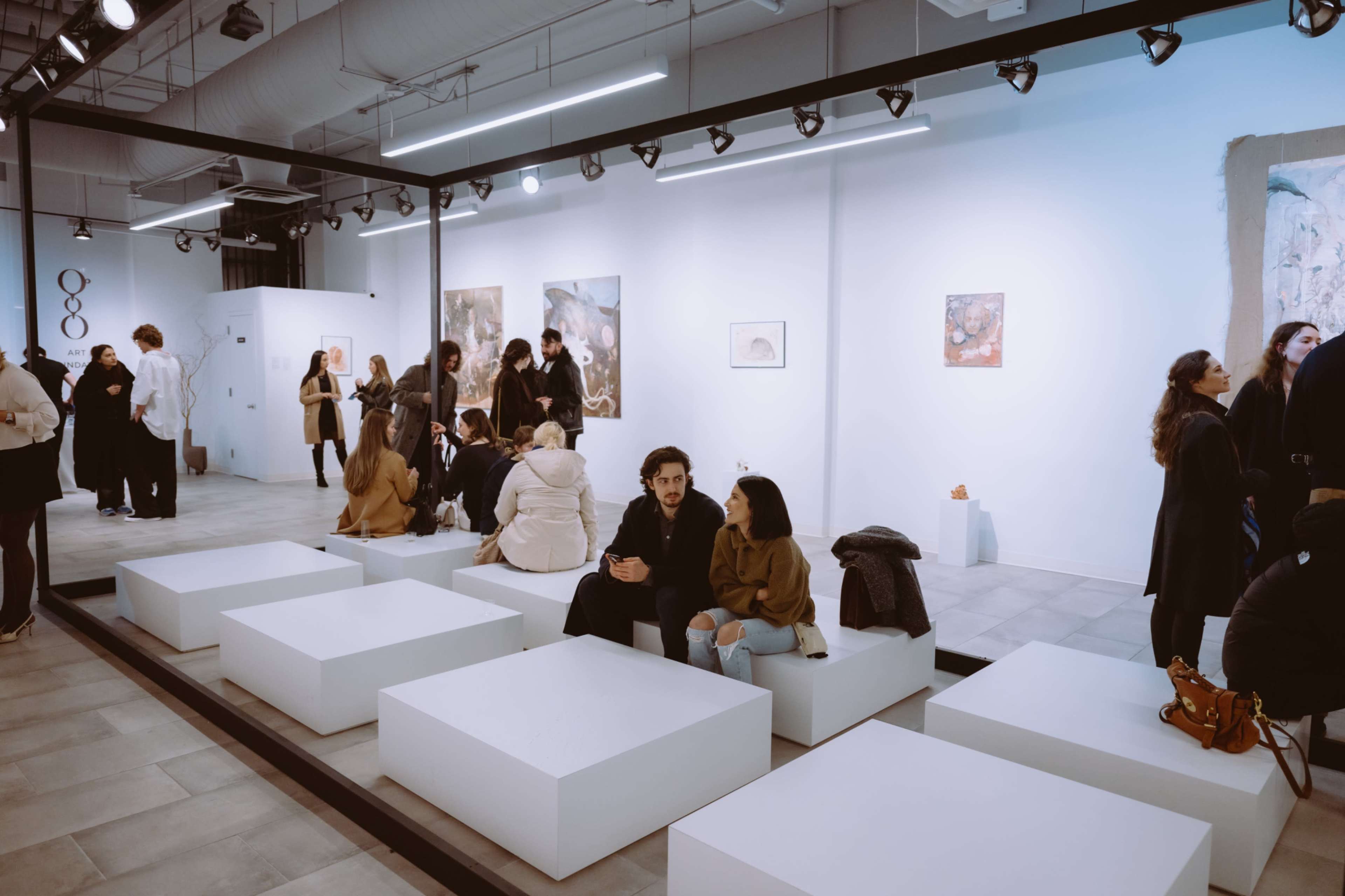 A crowded art gallery features guests conversing and viewing contemporary artwork on white walls, with minimalistic seating in the foreground.