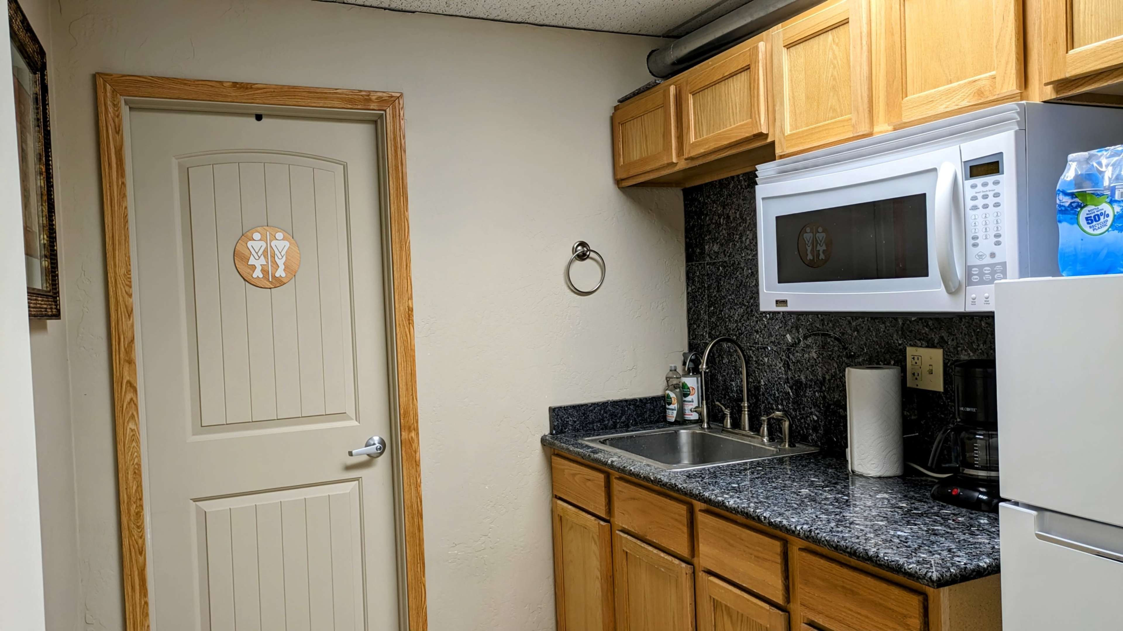 A small kitchen area with wooden cabinets, a sink, a microwave, and a closed door featuring a decorative emblem.