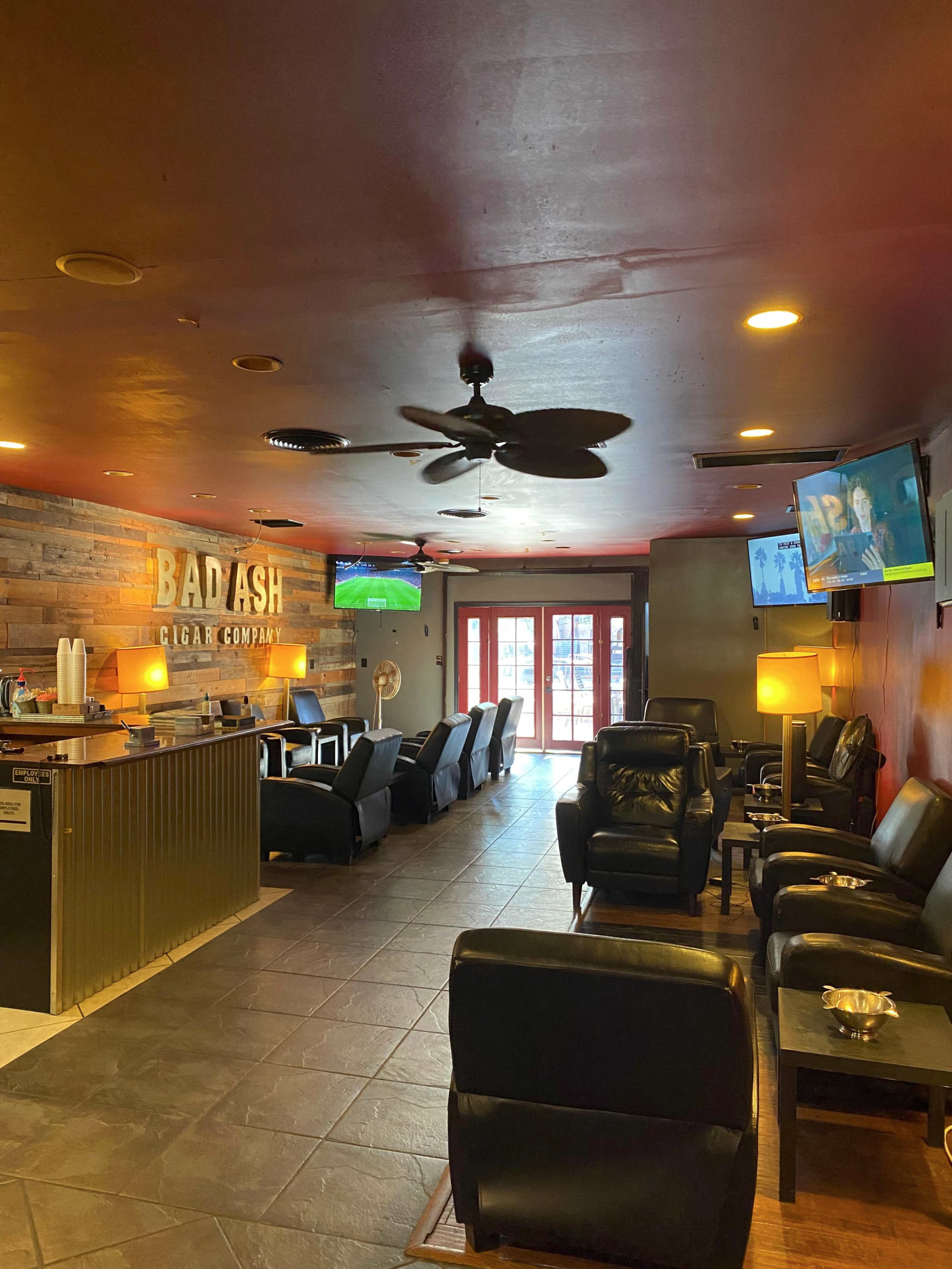 The interior of a cigar lounge features leather seating, a bar area, and televisions displaying sports.