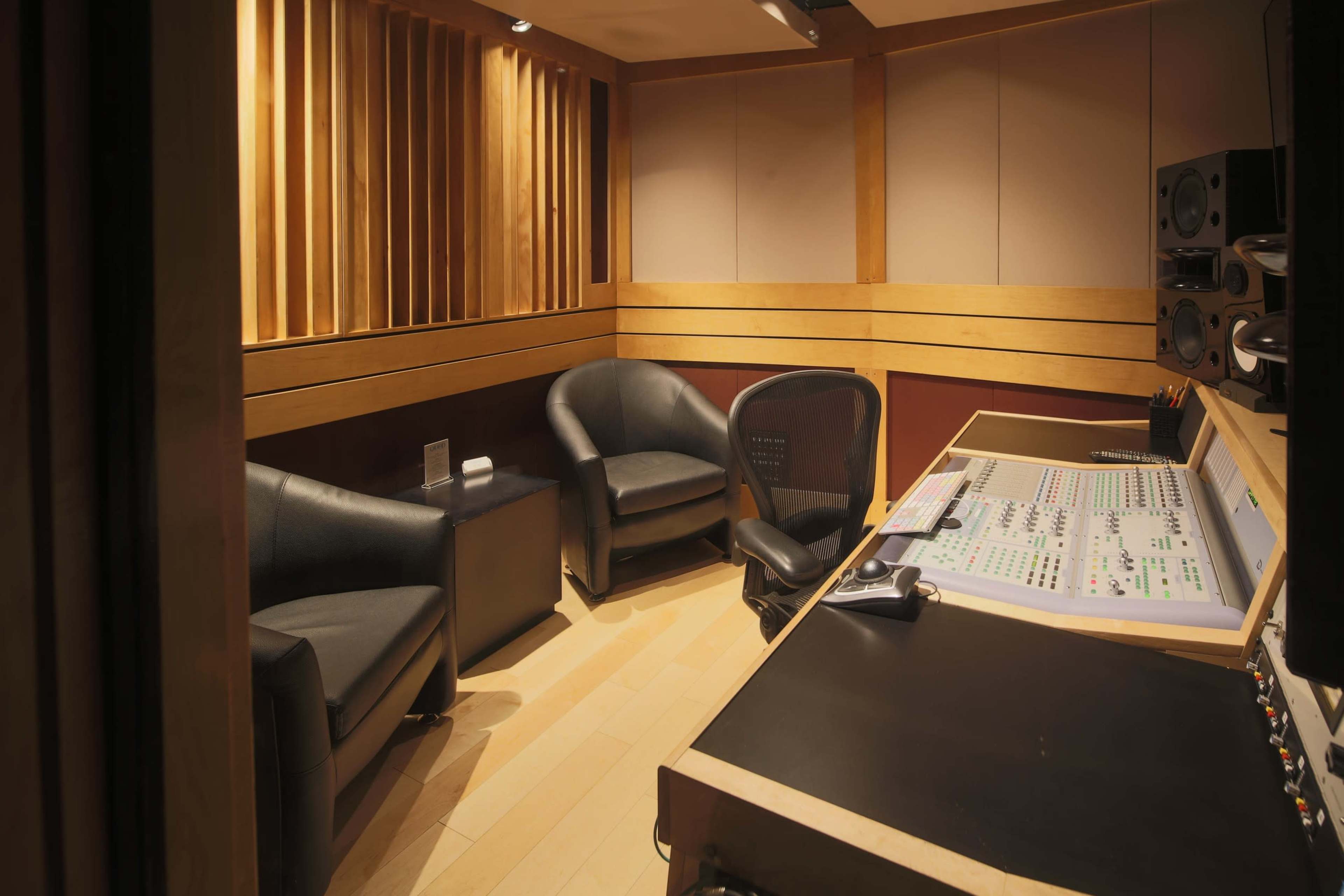 A sound recording studio features two black chairs and a control desk with a mixing console against wooden paneling.