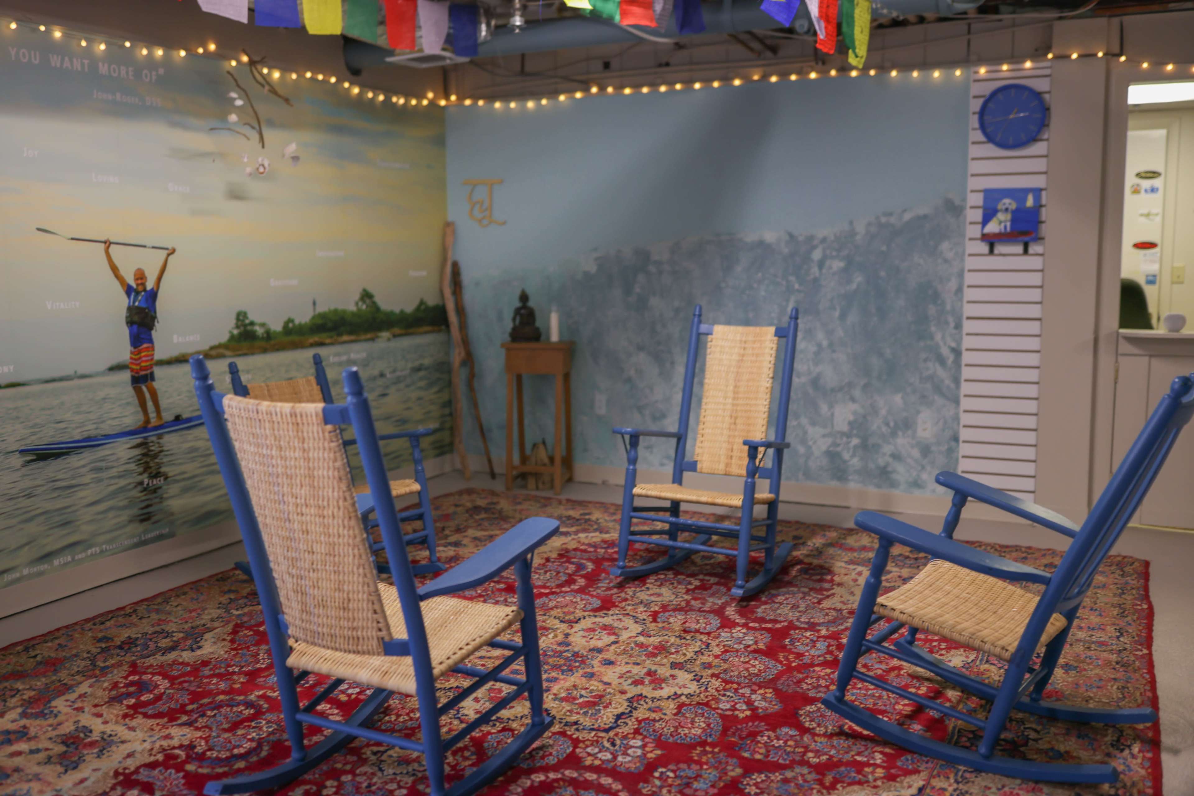 The image shows a room with four blue rocking chairs arranged in a circle on a patterned rug, against a wall featuring a mural of a person paddleboarding.