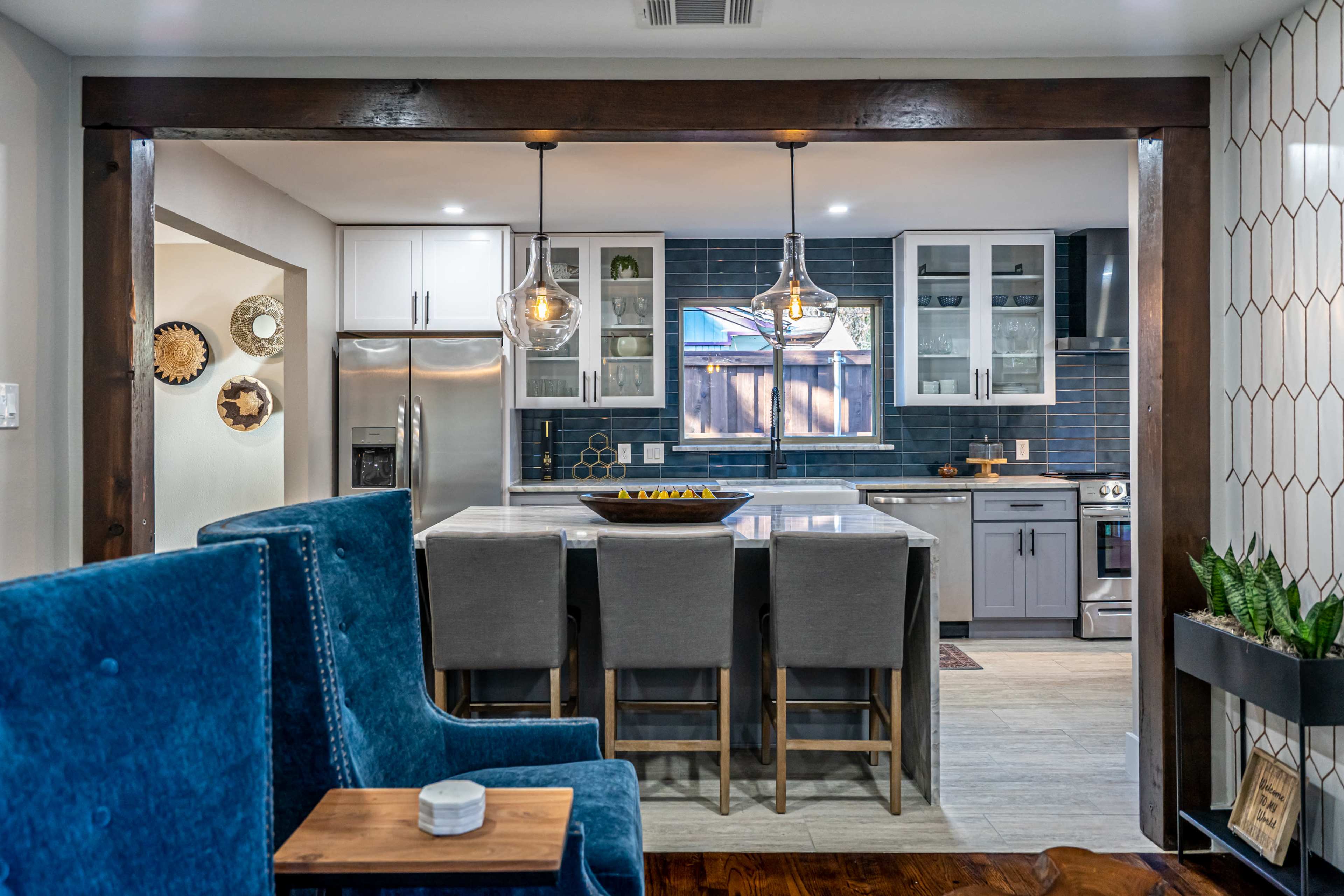 A modern kitchen is visible through an open entryway, featuring gray cabinetry, a large island with bar seating, and pendant lighting.
