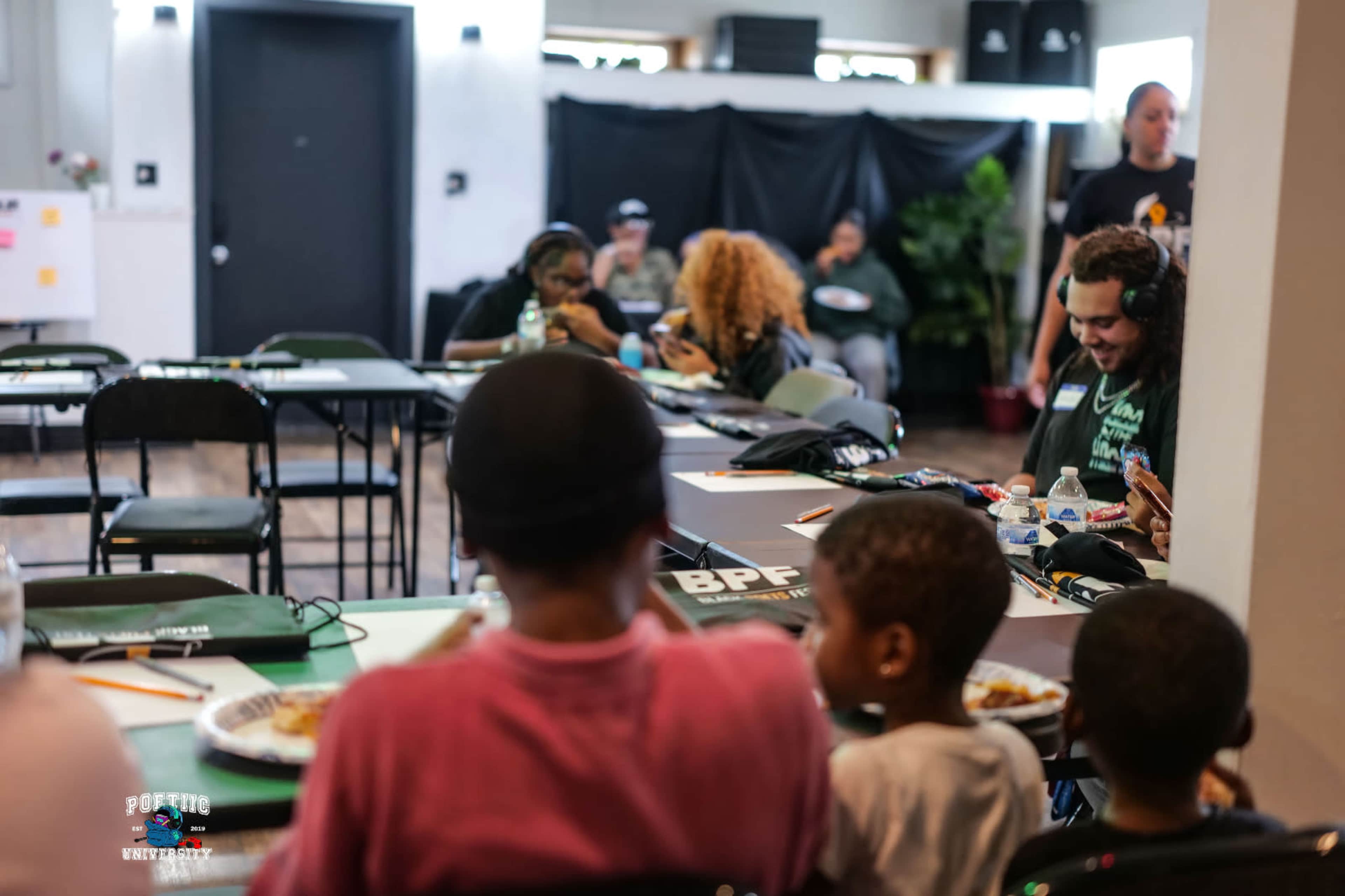 A group of children and adults are seated at tables in a classroom setting, engaged in various activities and eating food.