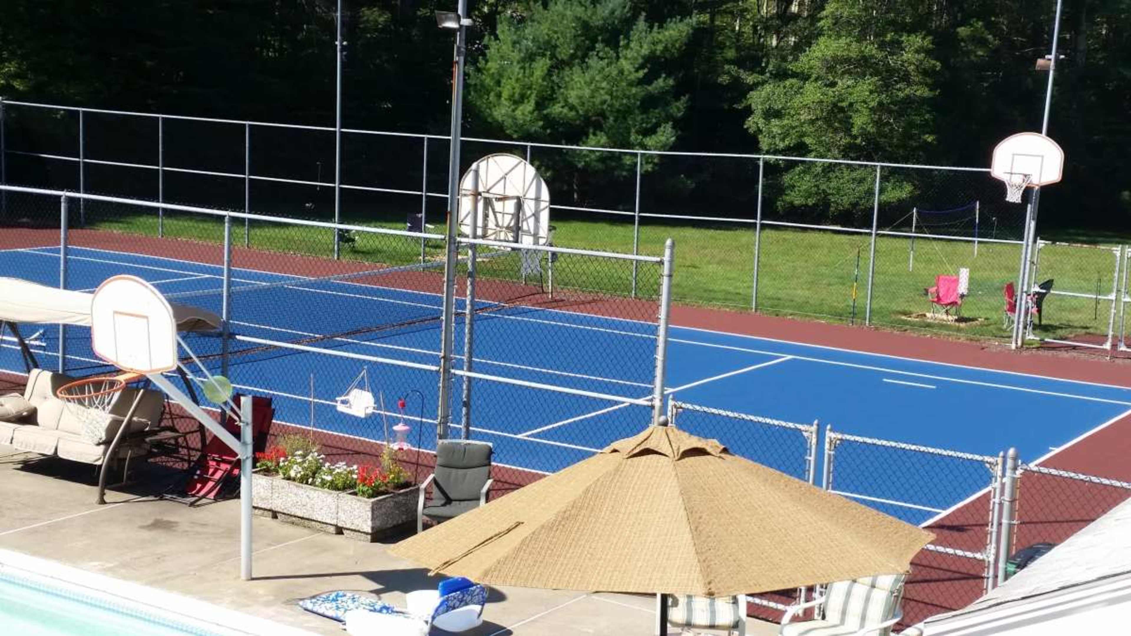 The image shows a multi-sport outdoor court with tennis and basketball areas, surrounded by a fence and landscaped with seating and umbrellas nearby.