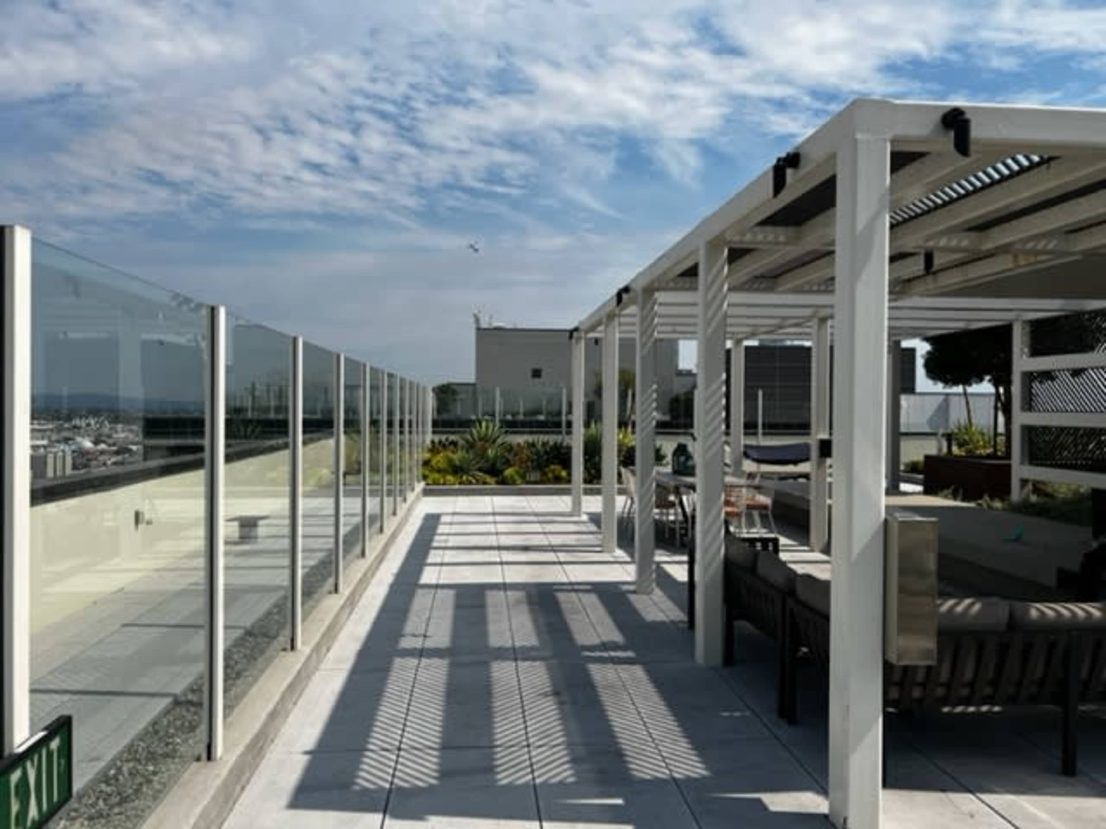 A rooftop patio with glass railings, potted plants, and seating arrangements under a partially cloudy sky.