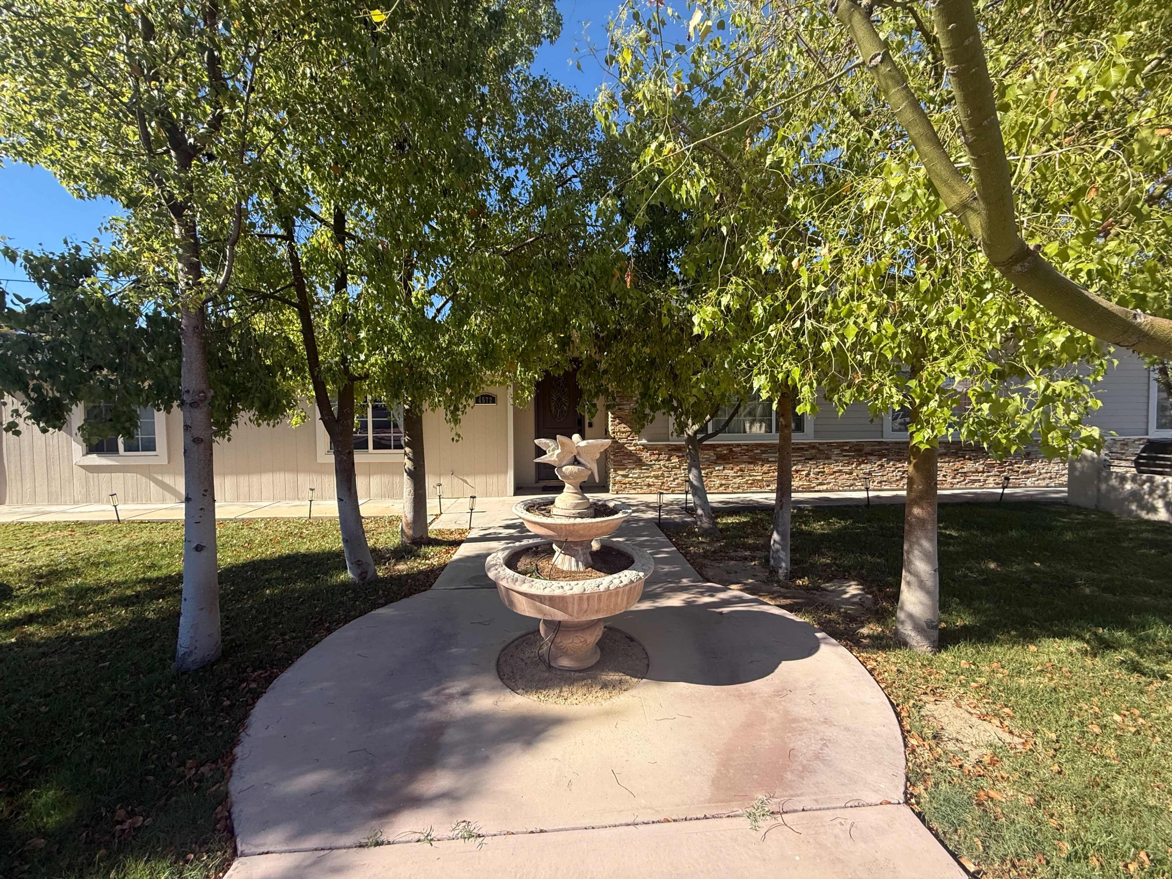 A stone fountain is positioned on a concrete pathway surrounded by trees leading to the entrance of a house.