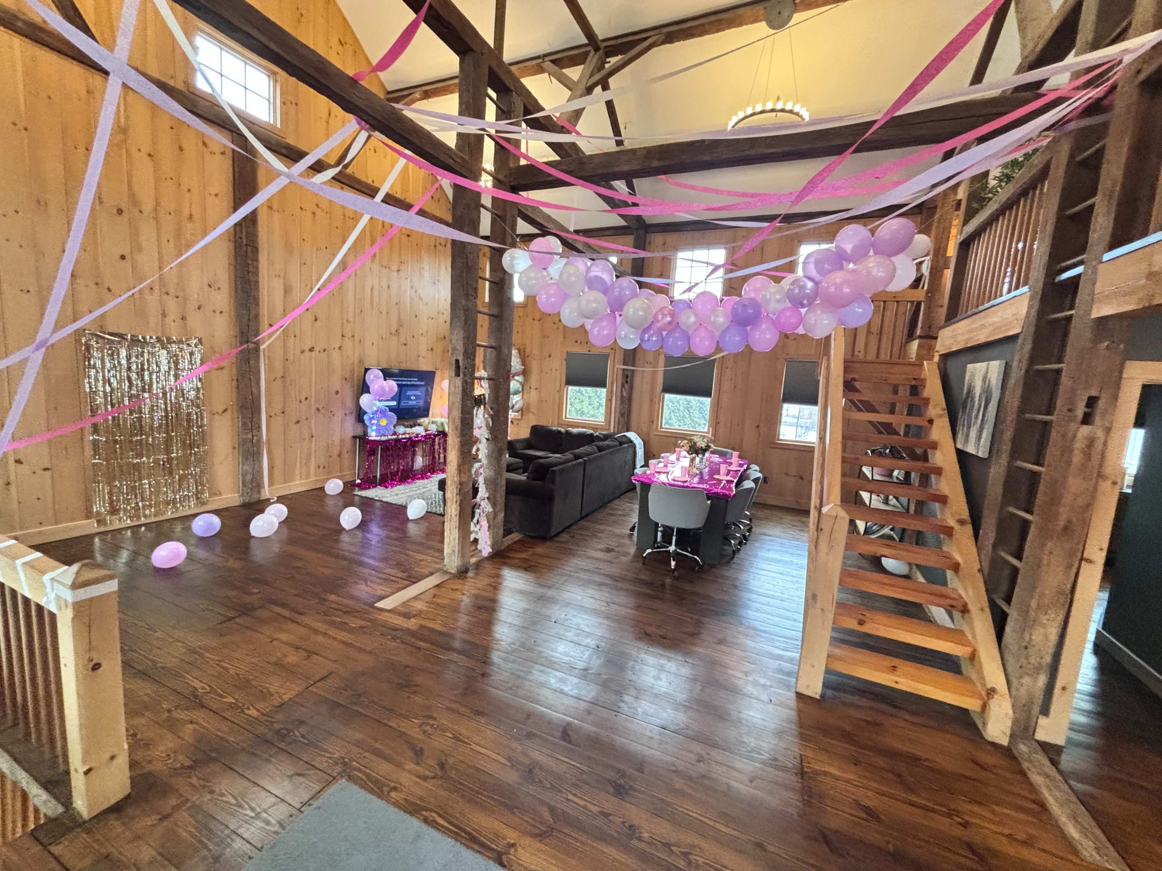 The image depicts a decorated indoor space featuring a table set for a celebration, with pink and purple balloons and streamers hanging from the ceiling, and a staircase leading to an upper level.