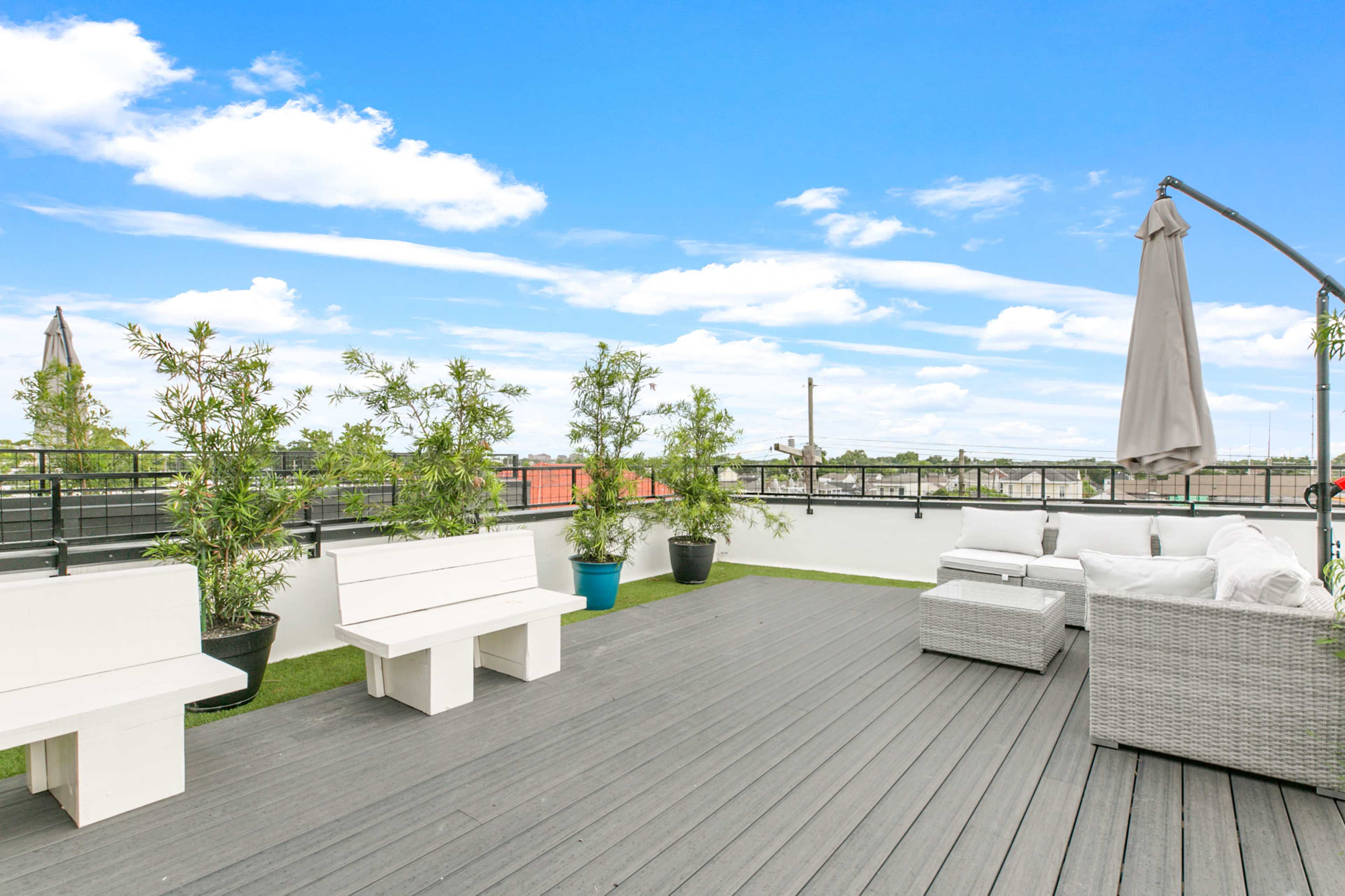A rooftop deck with white benches, potted plants, and a seating area surrounded by a clear blue sky.