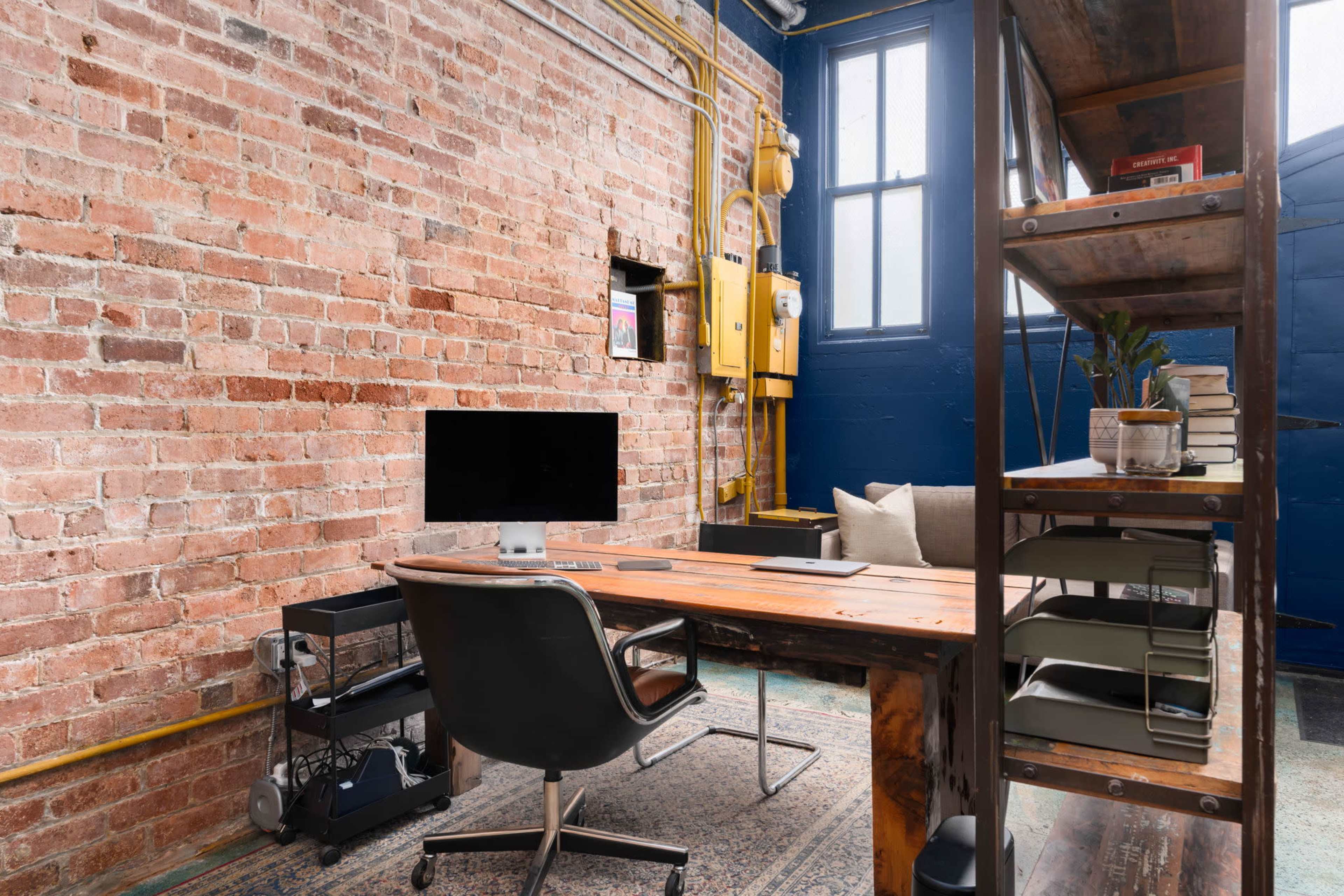 A small office space with a wooden desk, a computer monitor, a chair, and a metal shelving unit against a brick wall.