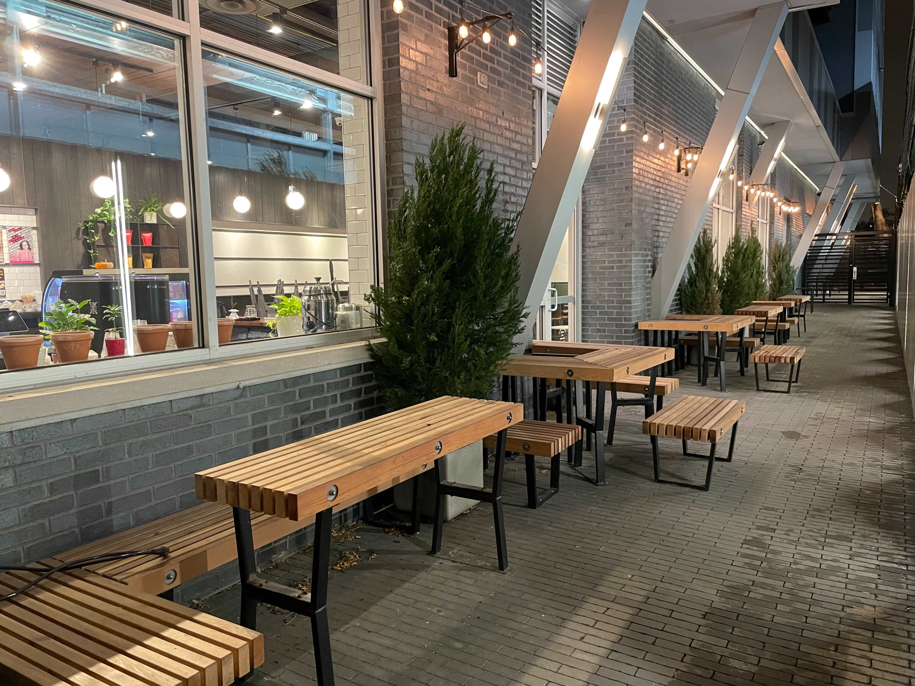The image shows a row of wooden tables lined up along a brick wall, with potted plants and bright lights above.