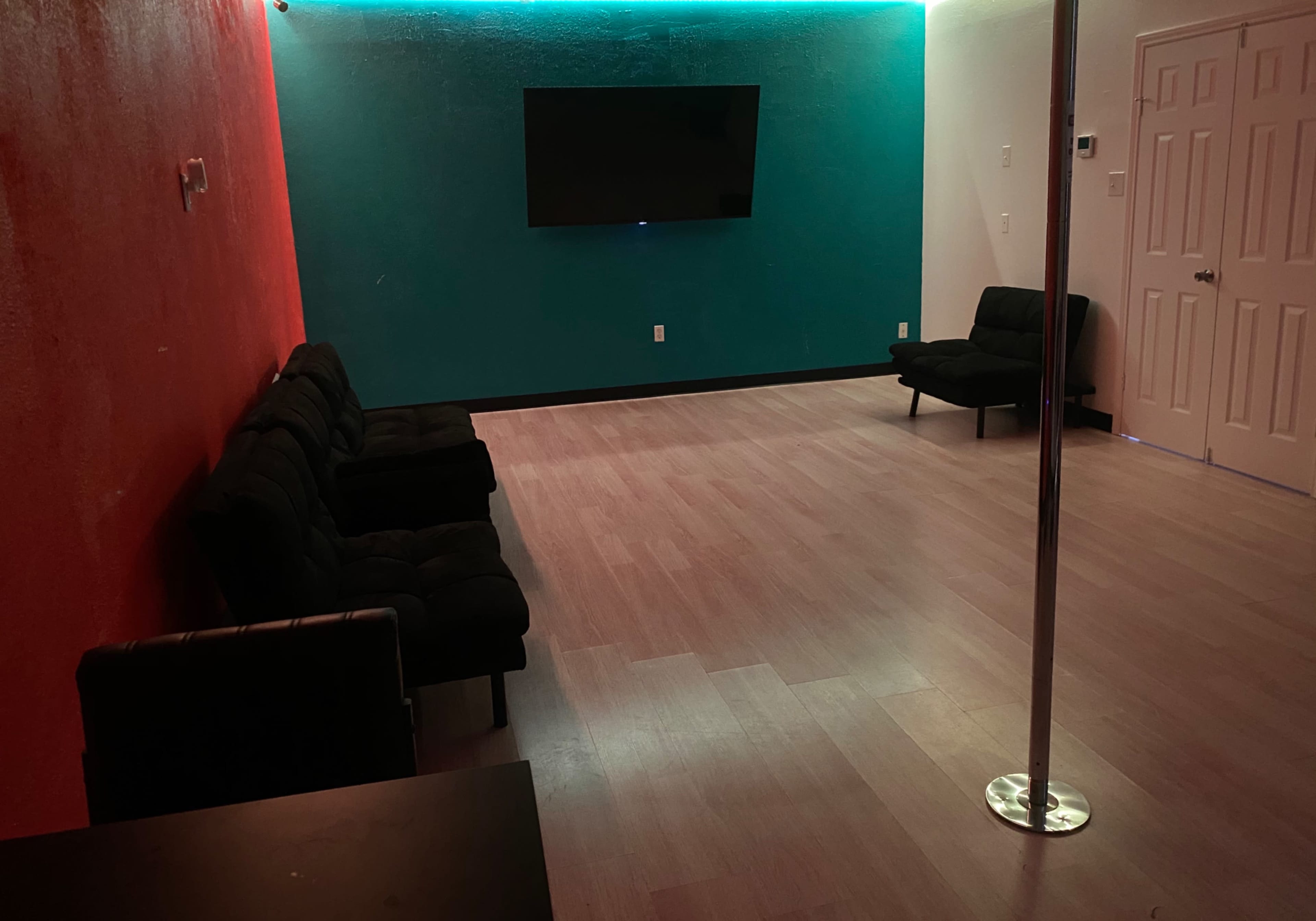 The room features hardwood flooring, a television mounted on a turquoise wall, a red accent wall, and seating arranged near a dance pole.