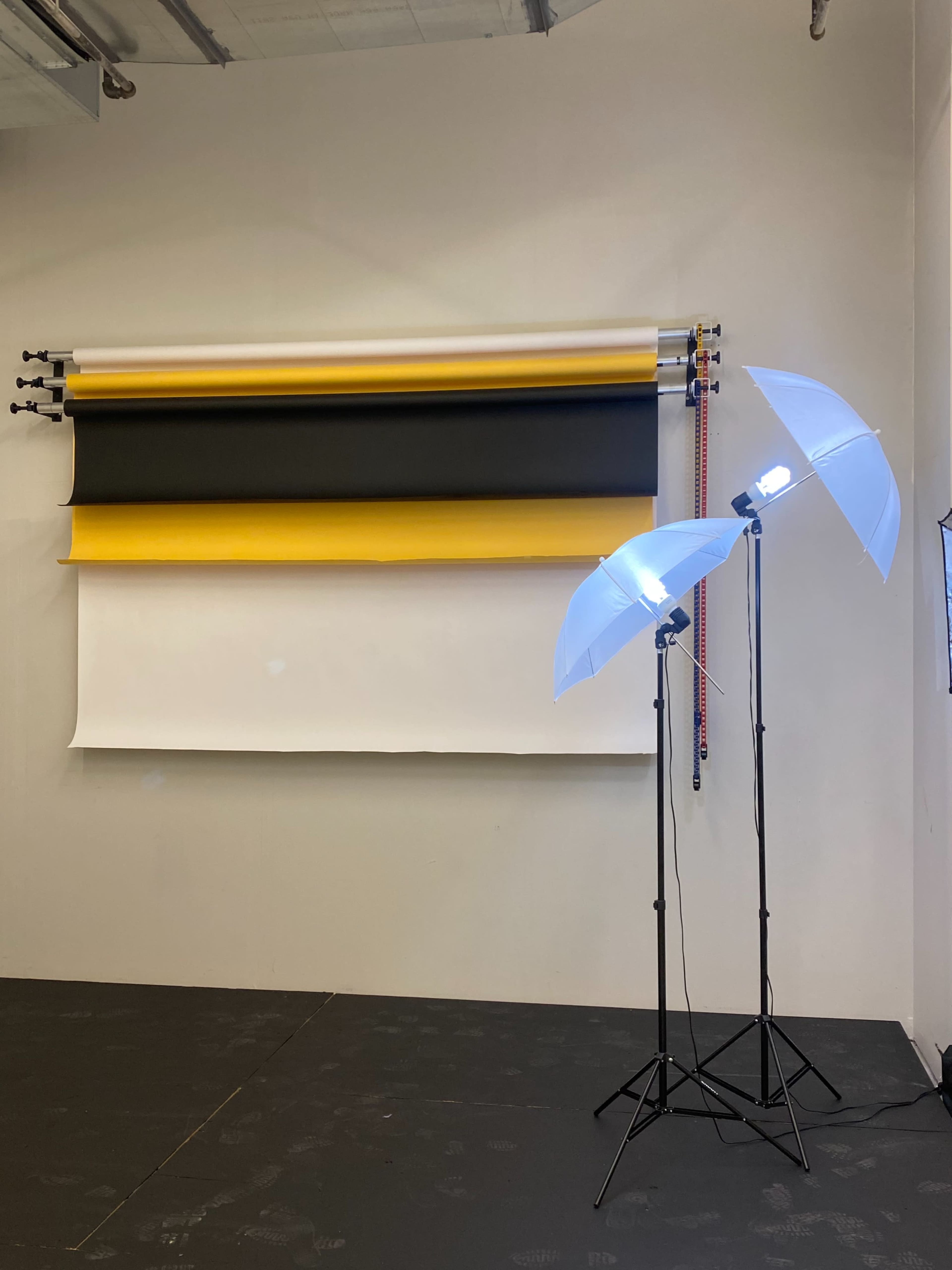 A photography studio setup with a backdrop consisting of yellow, black, and white rolls and two light stands with umbrellas positioned in front.