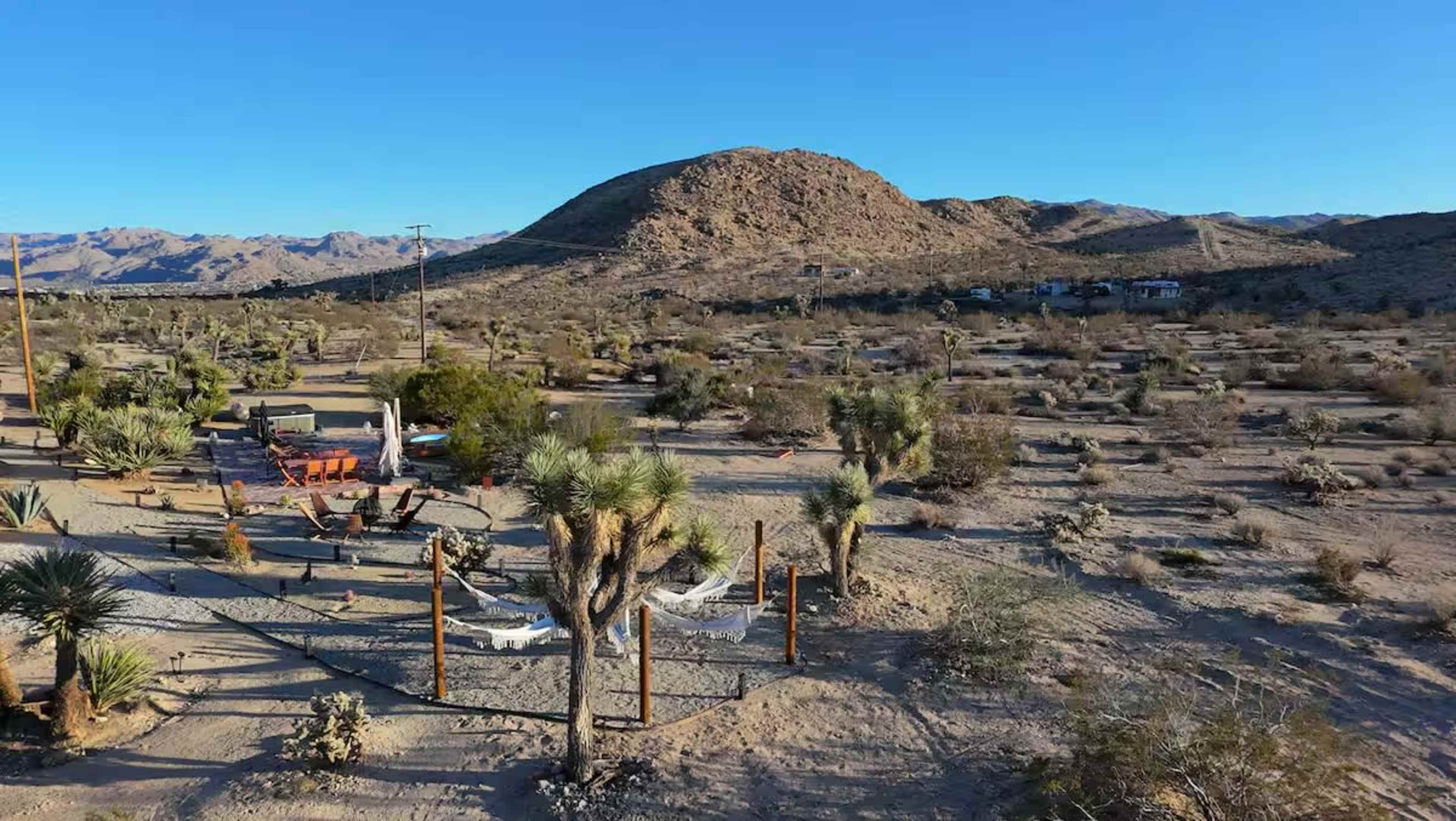 The image shows a desert landscape with sparse vegetation, rocky hills in the background, and a few structures scattered throughout the area.