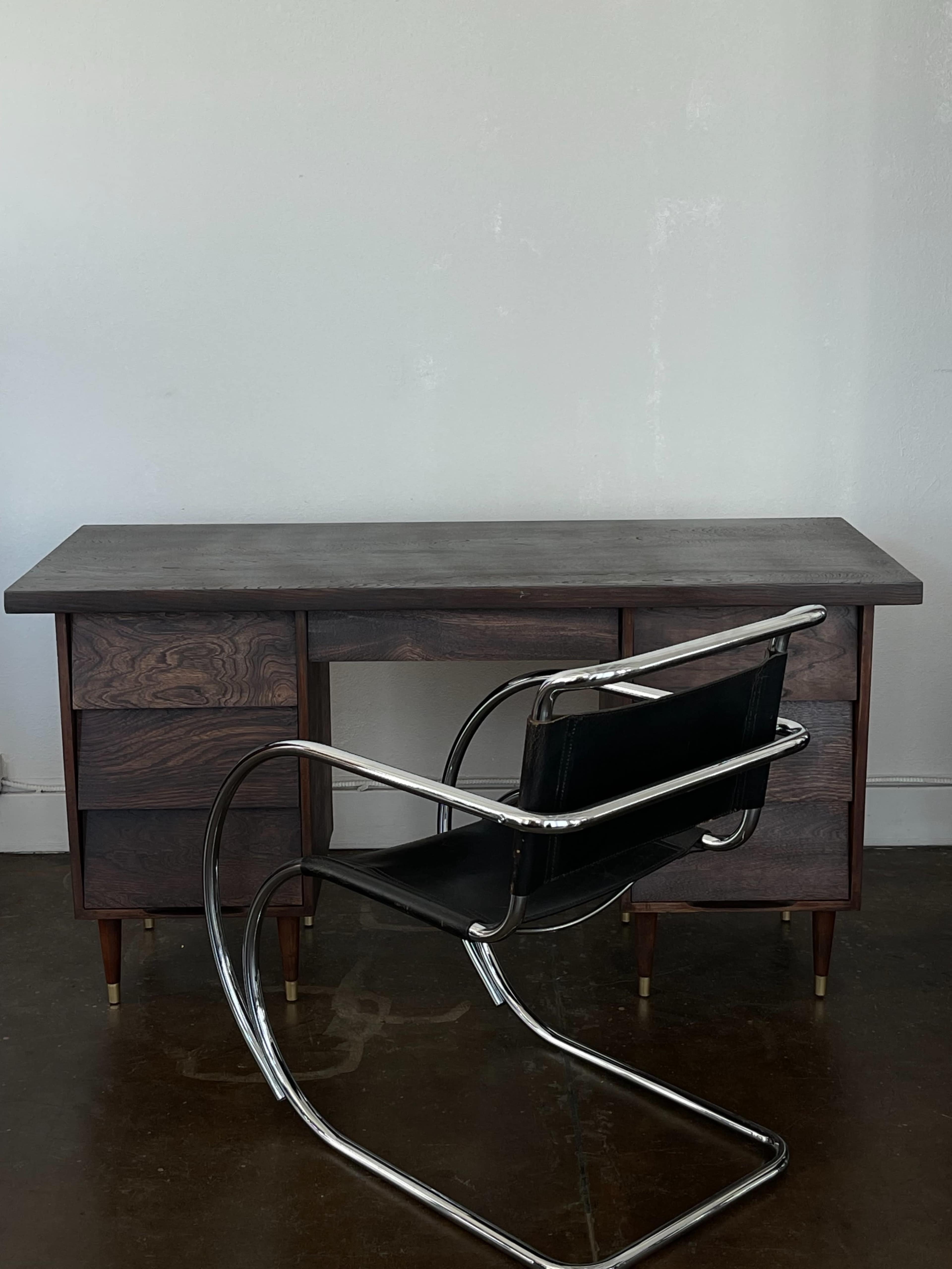 A wooden desk with two drawers and a modern metal chair stands in a minimalist room.