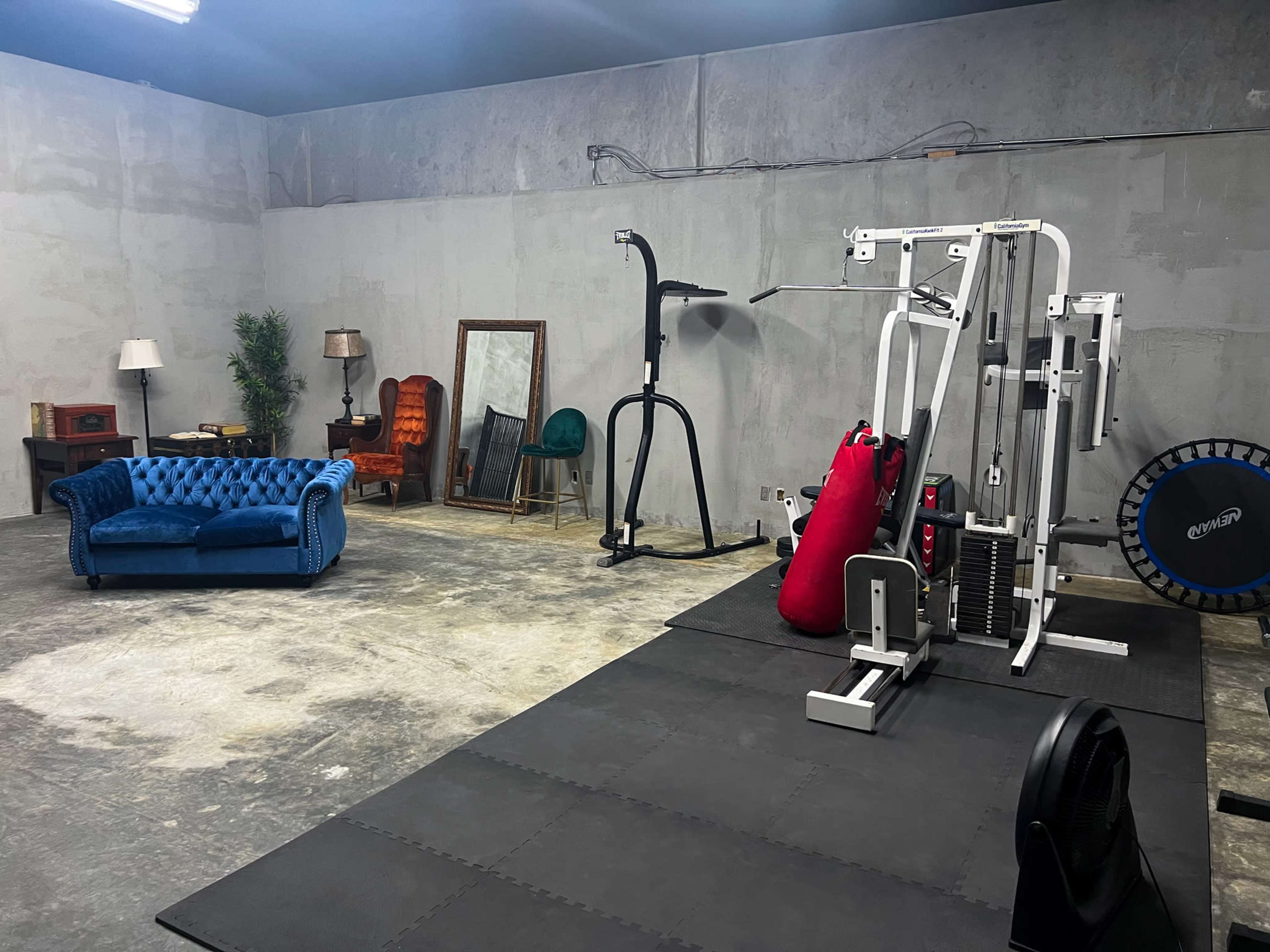An industrial-style gym interior with workout equipment on a black mat, a blue couch, a mirror, and various pieces of furniture against a concrete wall.