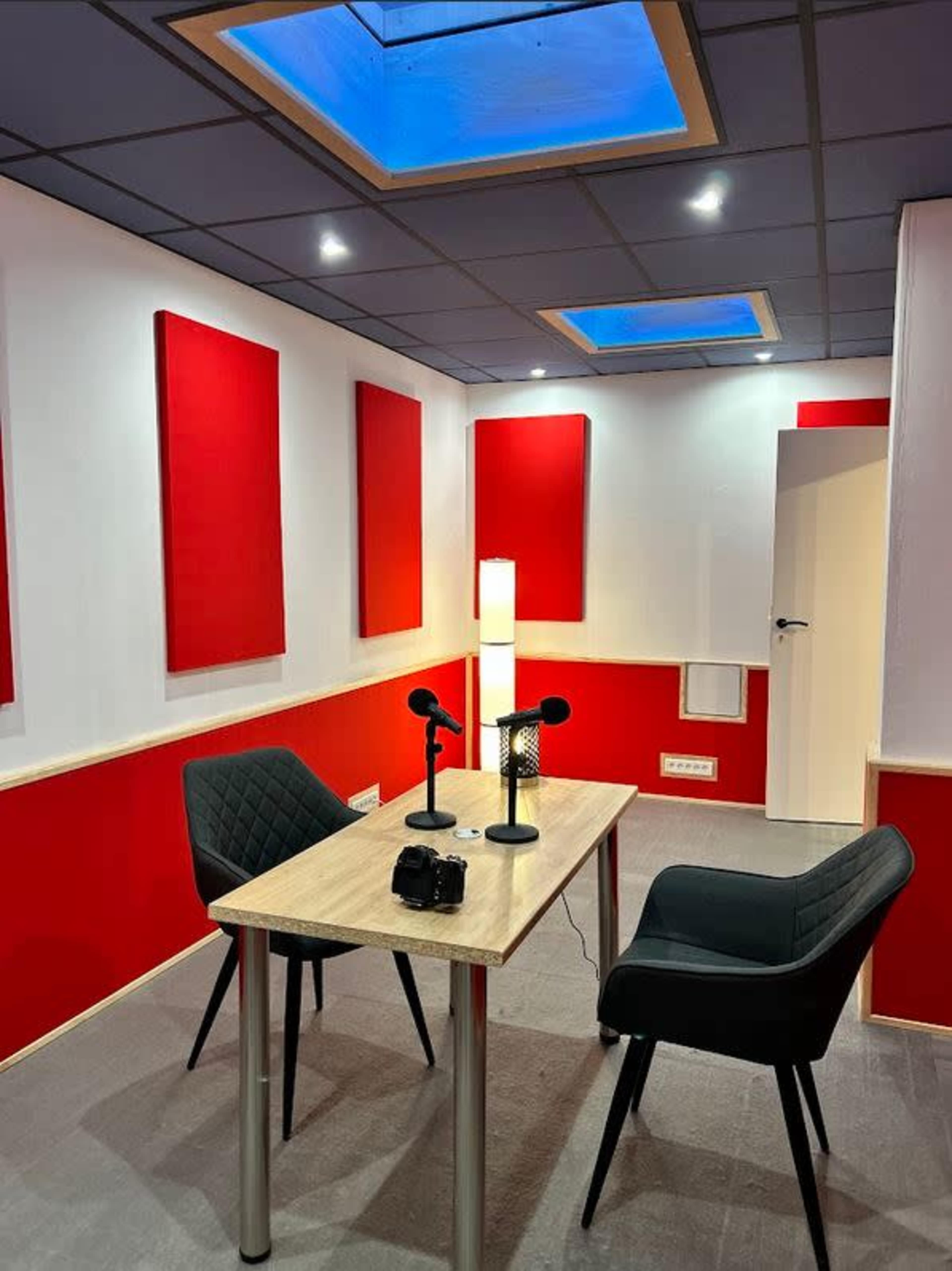 A small recording studio featuring a table with two microphones, two chairs, and red acoustic panels on the walls.