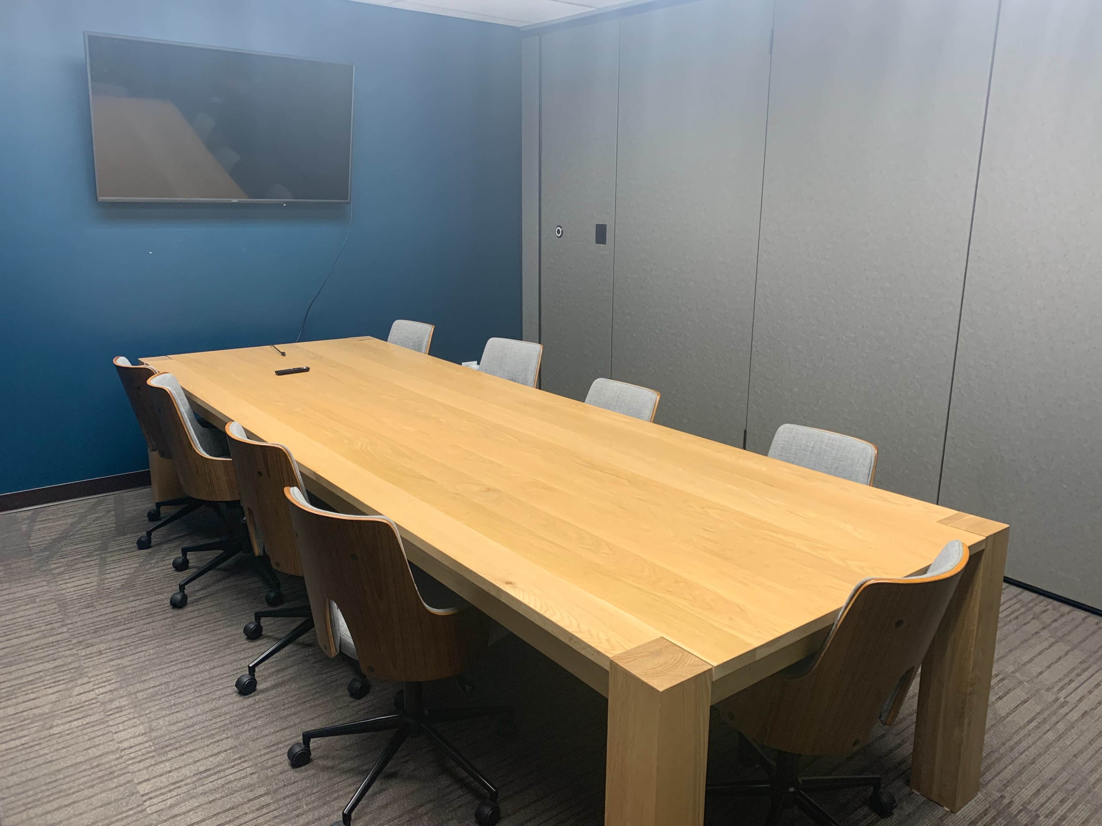 A large wooden conference table is surrounded by six ergonomic chairs in a meeting room with a wall-mounted television and a blue accent wall.