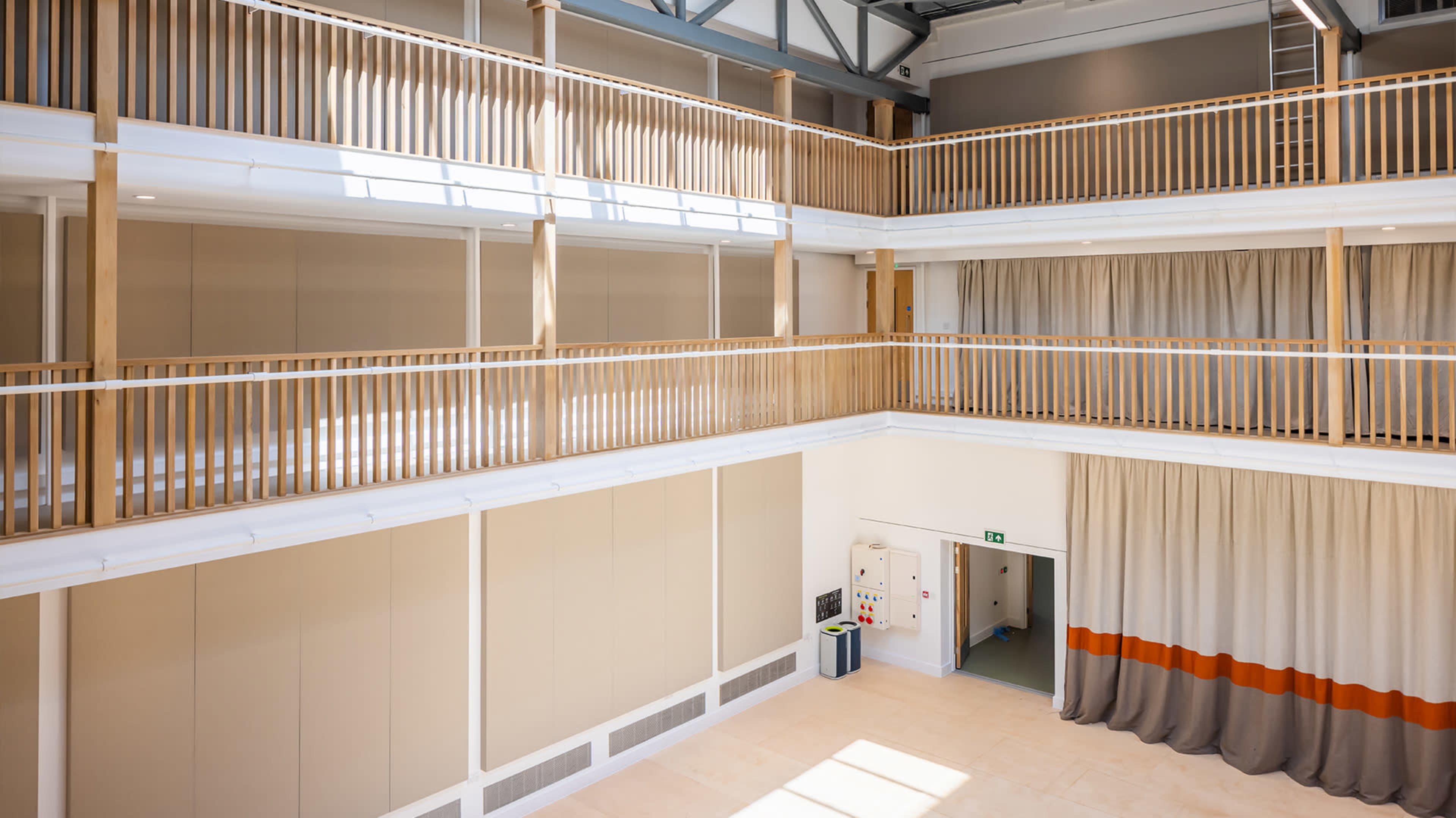 A spacious multi-level interior with wooden railings, beige walls, and large curtains, illuminated by natural light from windows above.