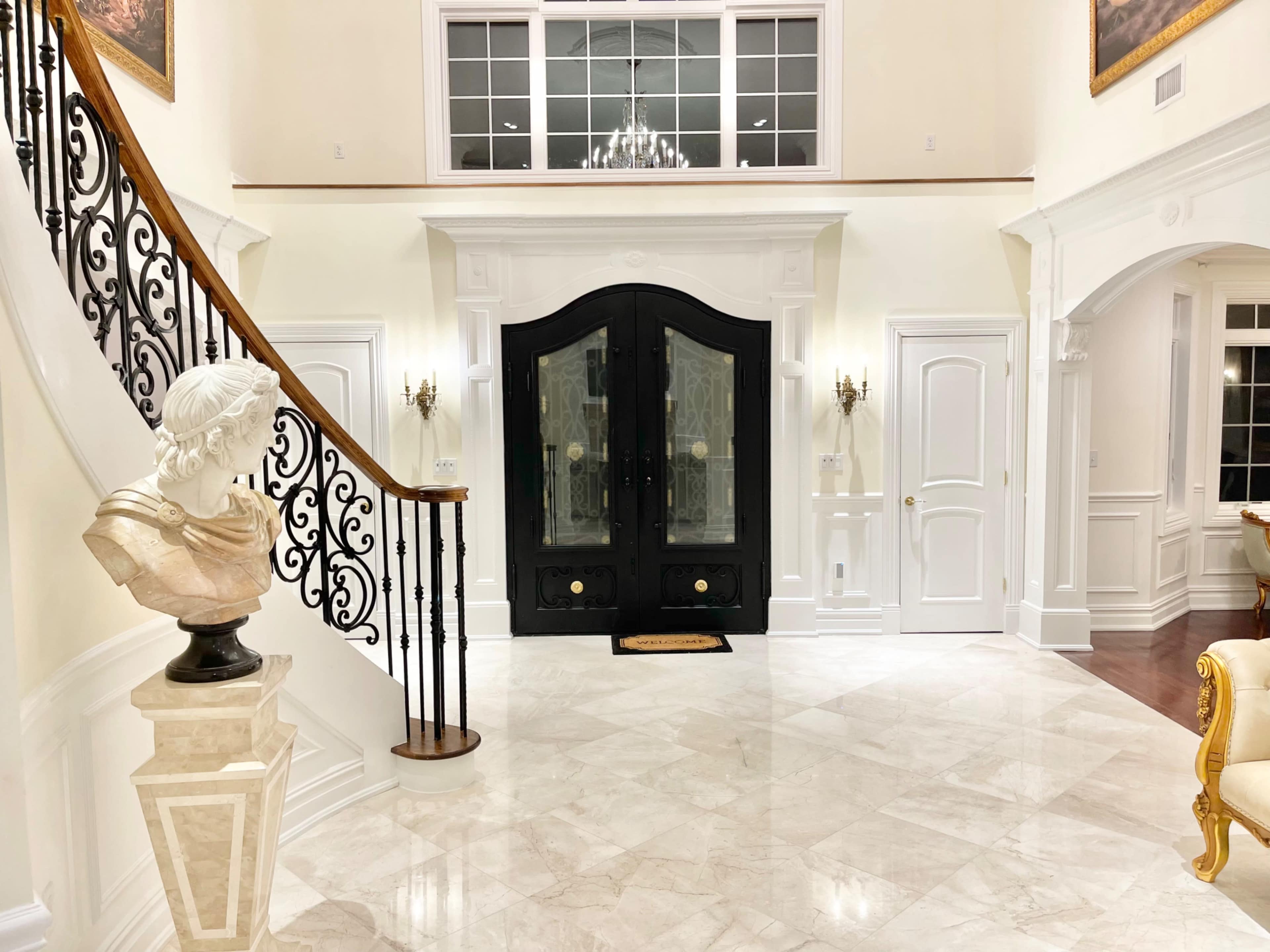 An elegant foyer with a spiral staircase, a bust on a pedestal, and large black double doors framed by white walls and marble flooring.