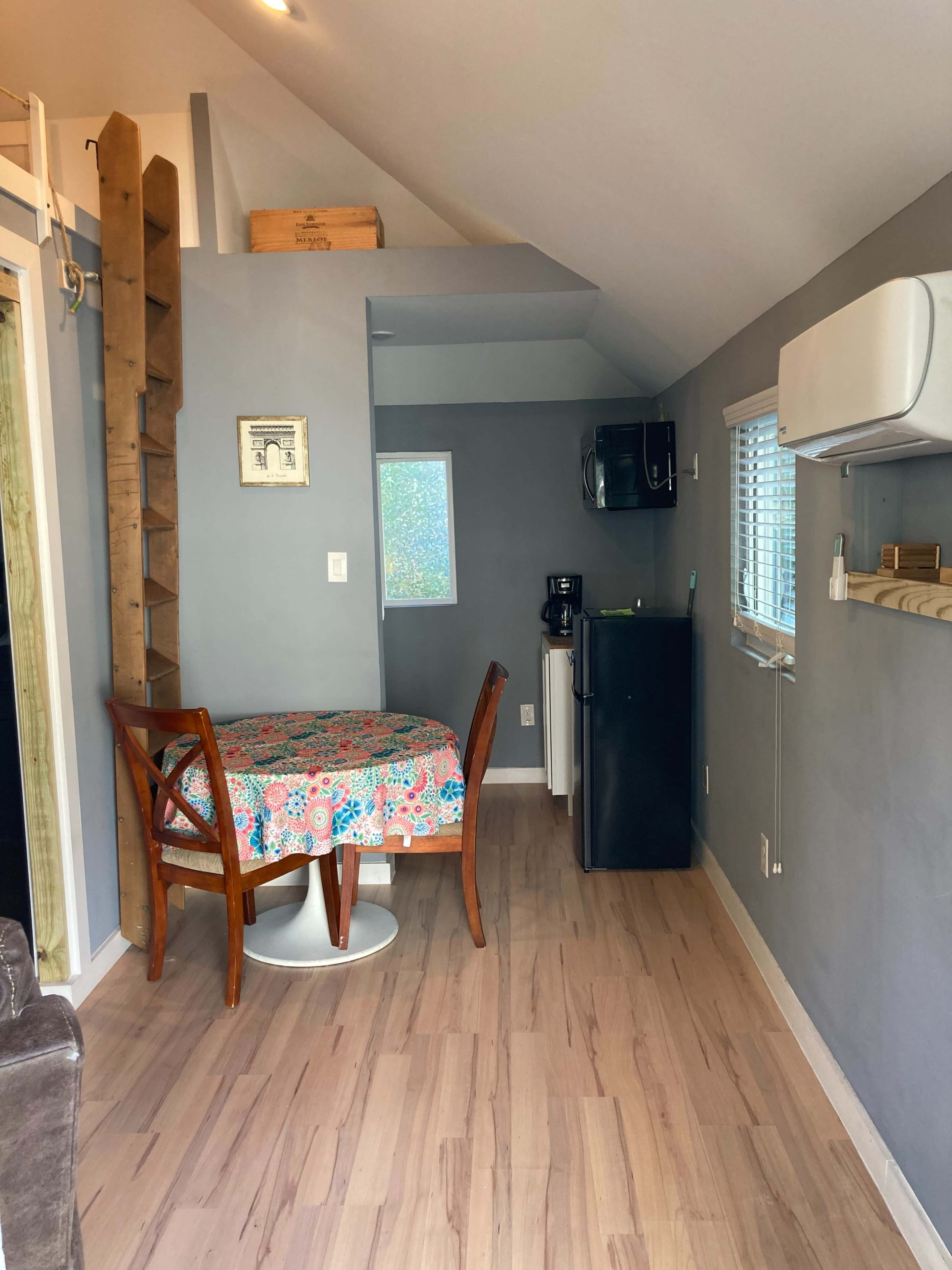 A small dining area with a round table covered in a floral tablecloth, a kitchenette with a fridge and coffee maker, and a wooden ladder leaning against the wall.
