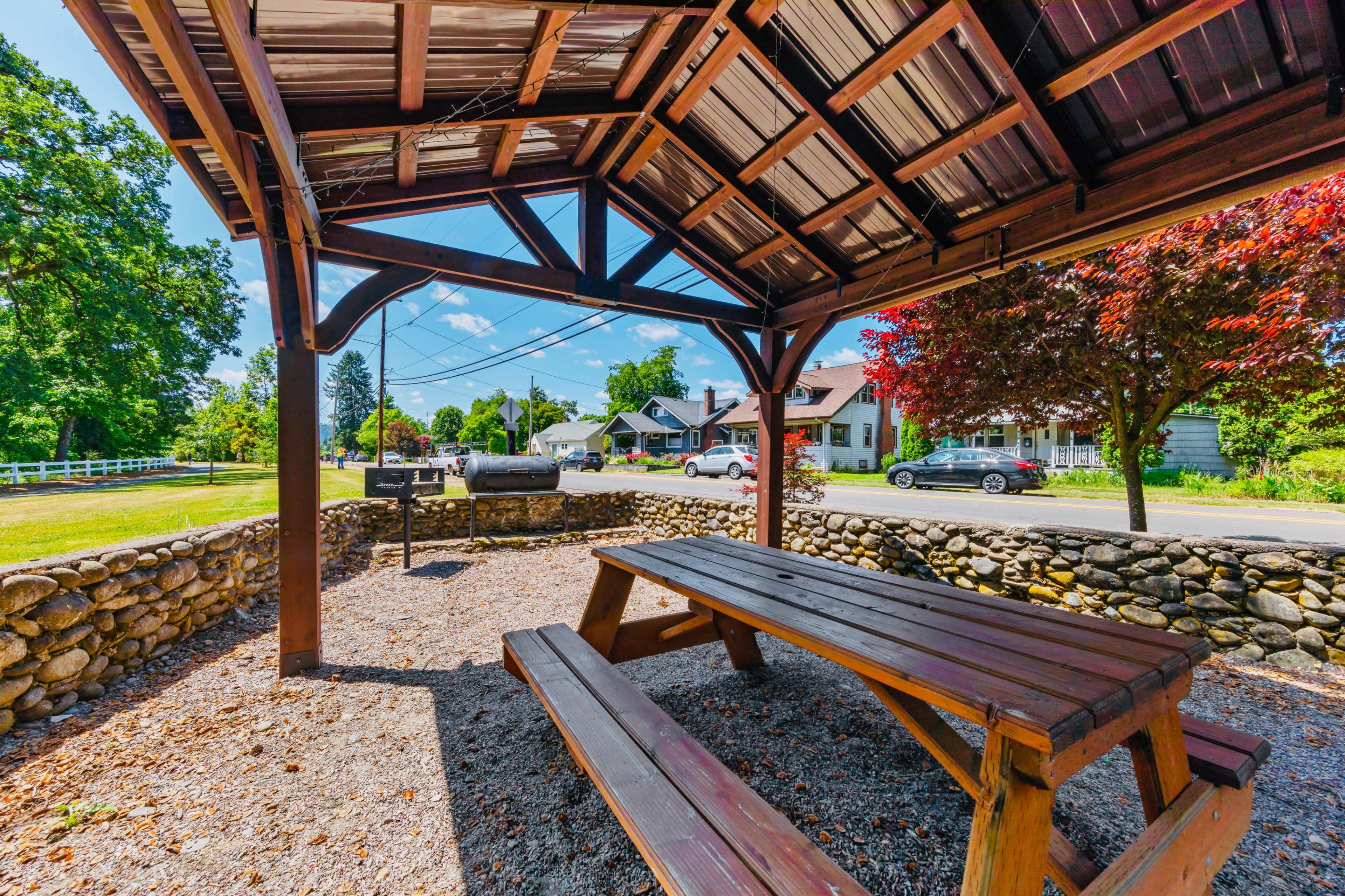 A covered picnic area with a wooden table and grill is positioned next to a stone wall, overlooking a residential street lined with trees and parked cars.
