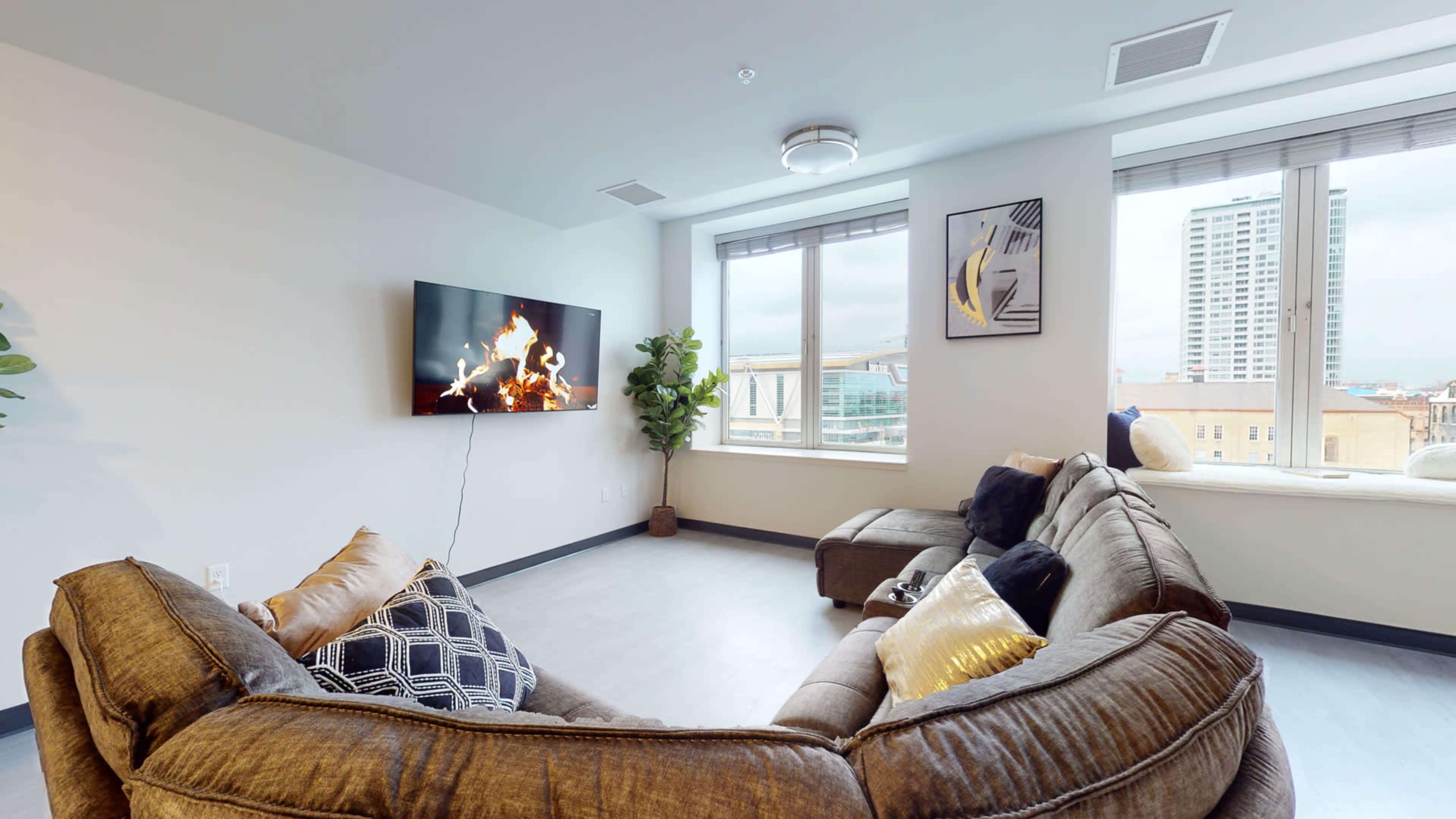 A modern living room features a large sectional sofa arranged around a wall-mounted television displaying a fire, with windows offering views of a cityscape.